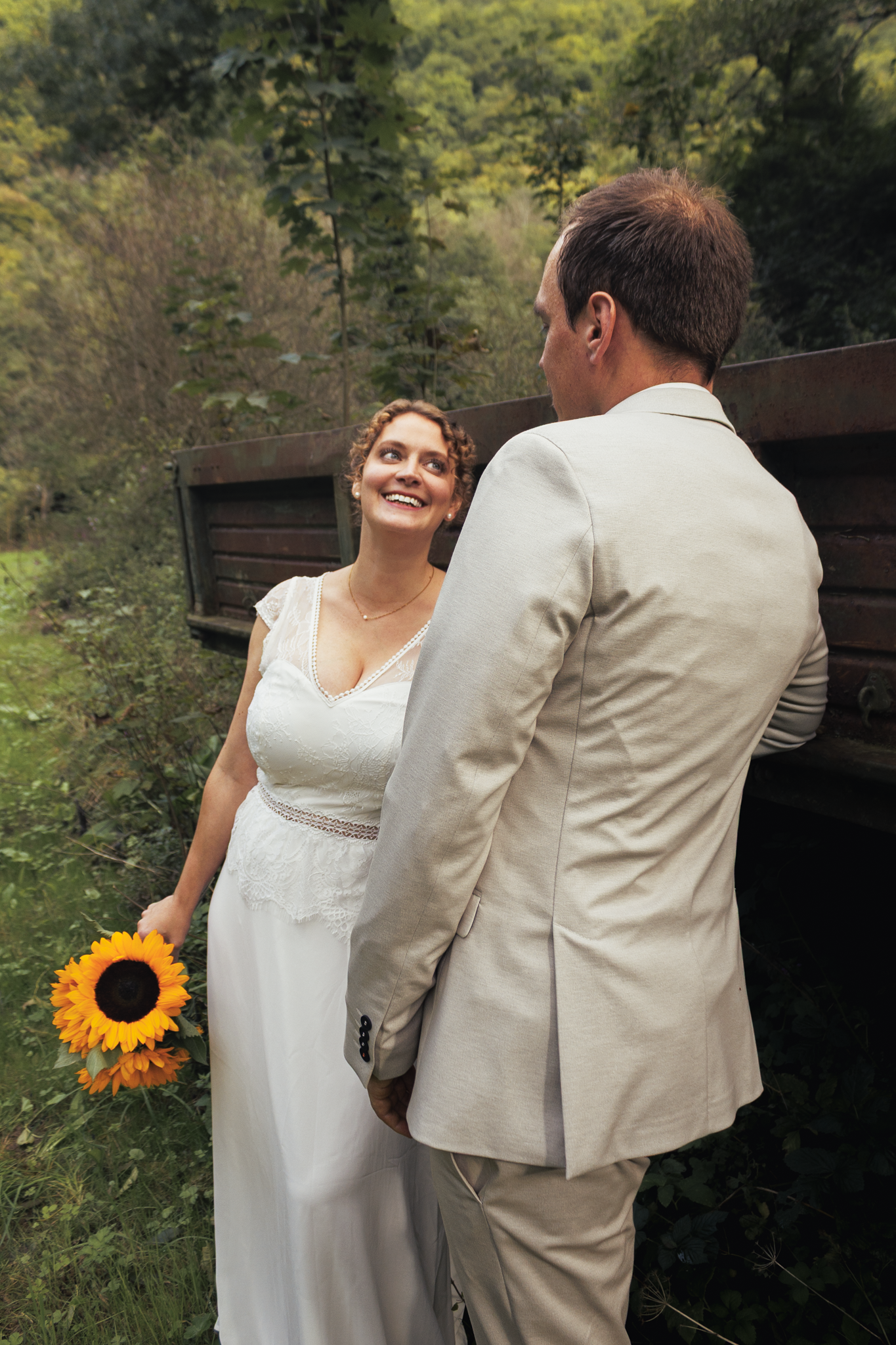 Ein Paar in formeller Kleidung, vermutlich frisch verheiratet, steht im Grünen. Die Frau trägt ein weißes Abendkleid und hält einen Sonnenblumenstrauß, während der Mann einen beigen Anzug trägt. Ein Lächeln verbunden mit Blickkontakt ist sichtbar, di