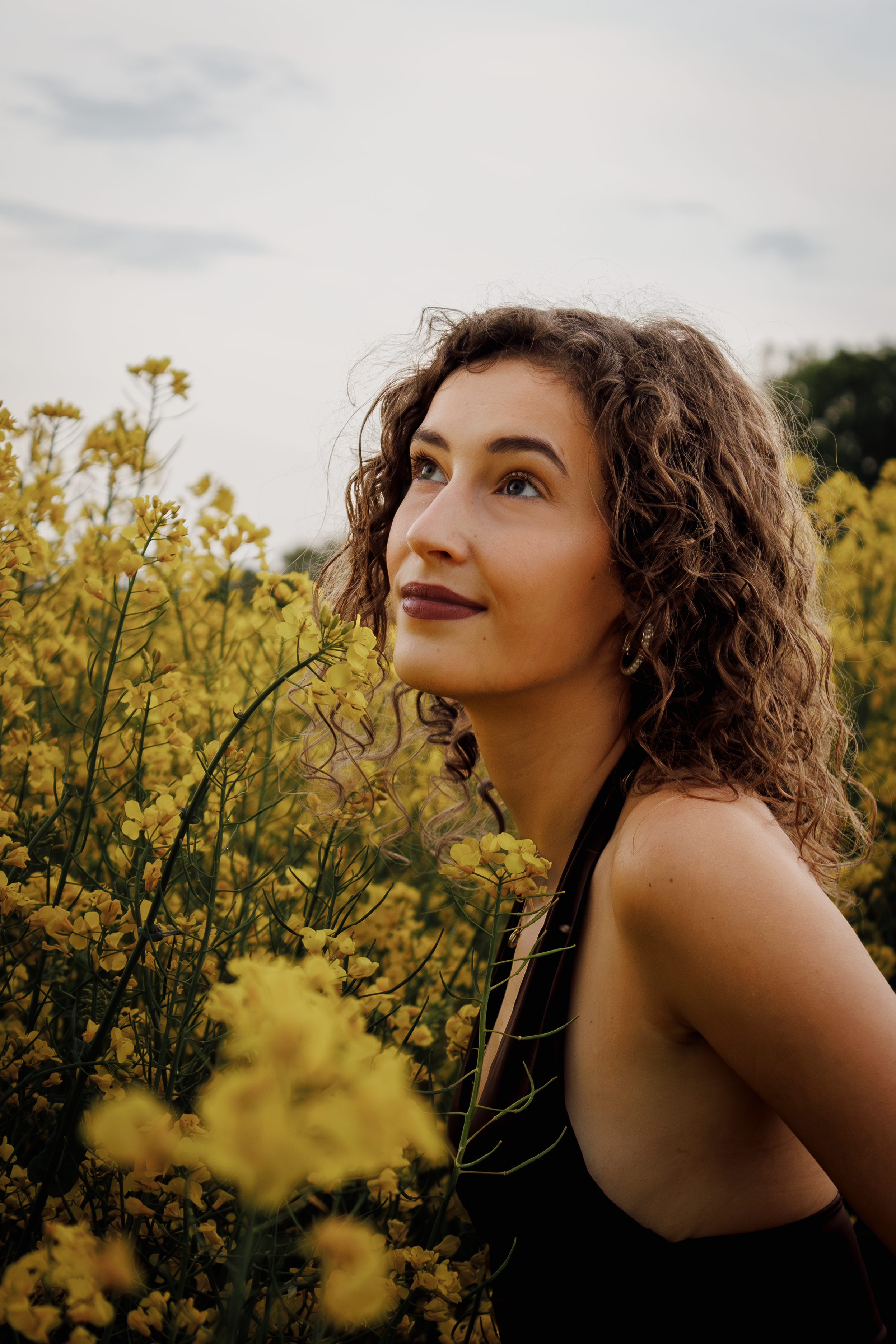 Junge Frau mit lockigem Haar, lächelnd, in einem schwarzen Kleid, inmitten gelber Blumen im Freien, Blick nach oben, bei leicht bewölktem Himmel.