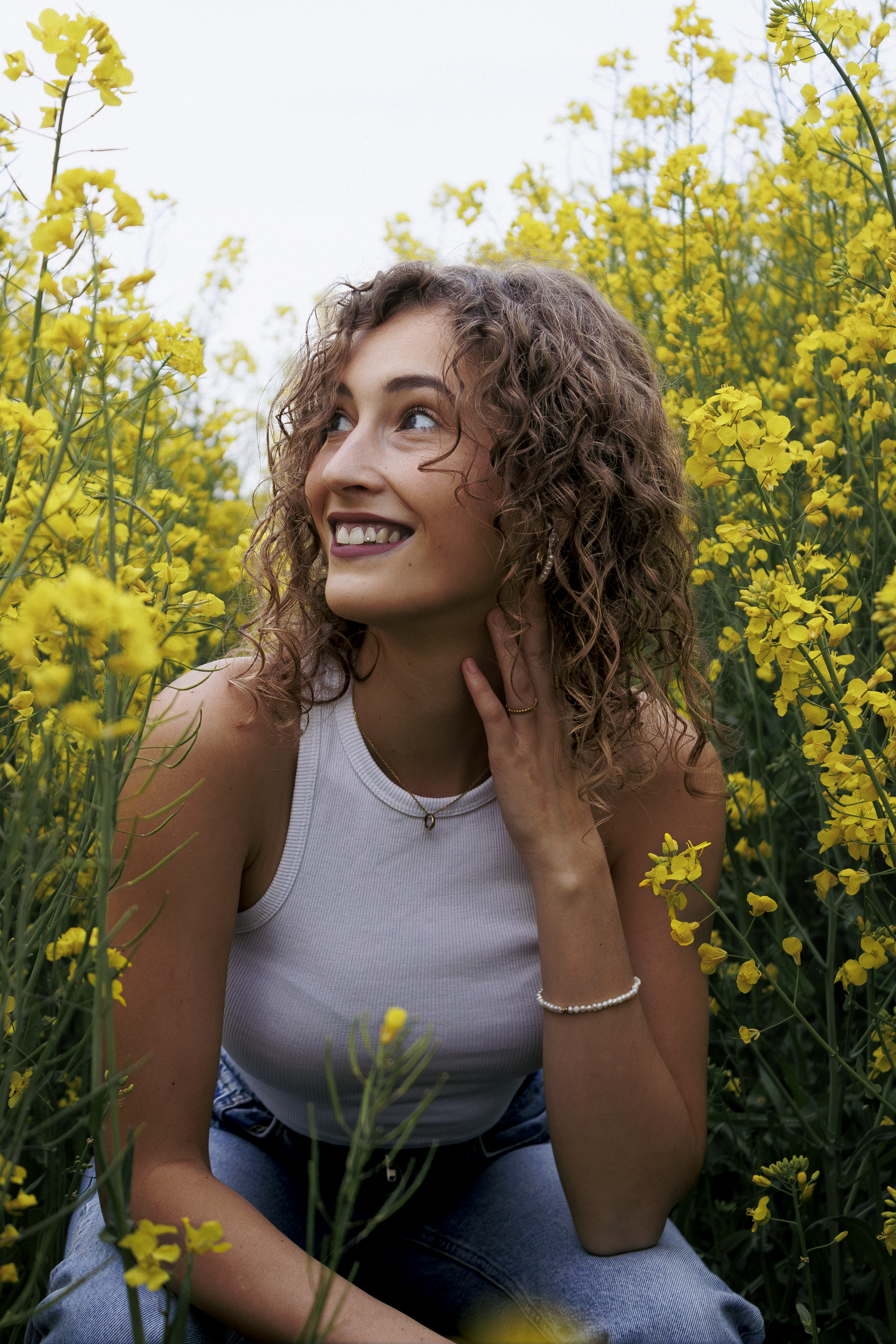 Junge Frau mit lockigem Haar, lächelnd und blickend nach oben, sitzend in einem gelben Blumenfeld, trägt ein weißes Tanktop, Jeans und Schmuck, umgeben von vielen gelben Blumen.