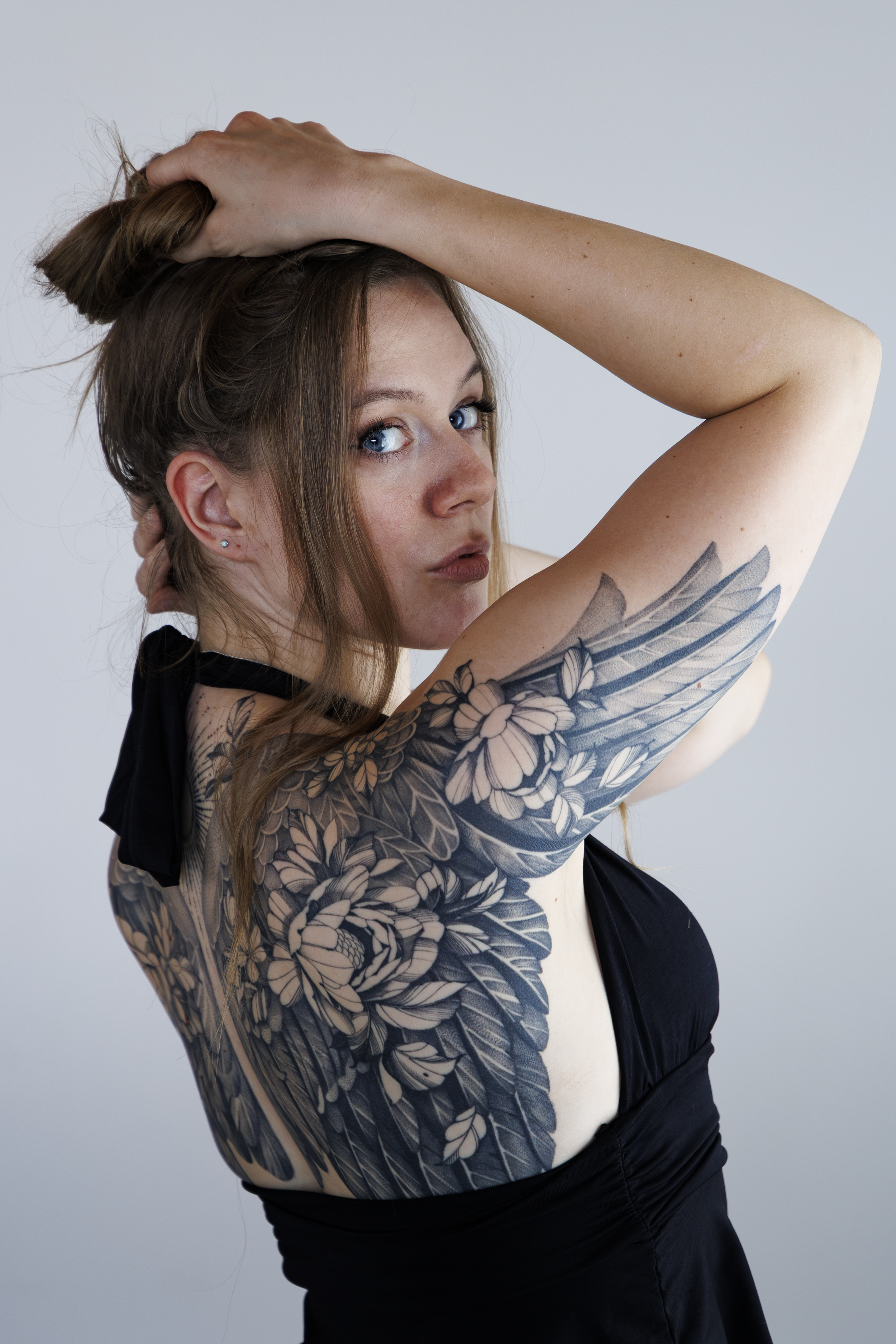 Junge Frau mit Tattoo auf dem Arm hebt die Arme und trägt schwarze Kleidung, blickt in die Kamera, vor hellem Hintergrund.