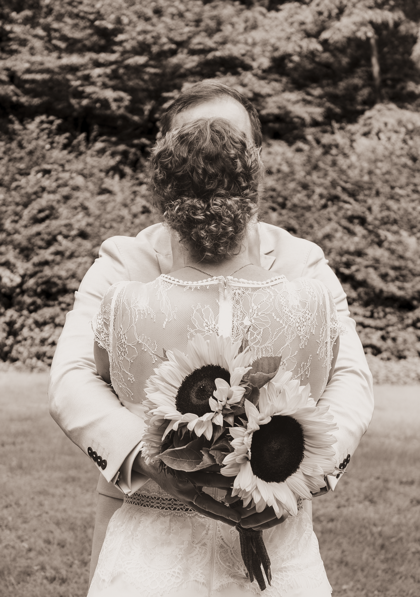 Ein Paar bei der Hochzeit, das sich umarmt, während die Braut einen Sonnenblumenstrauß hält, im Freien mit Bäumen im Hintergrund.