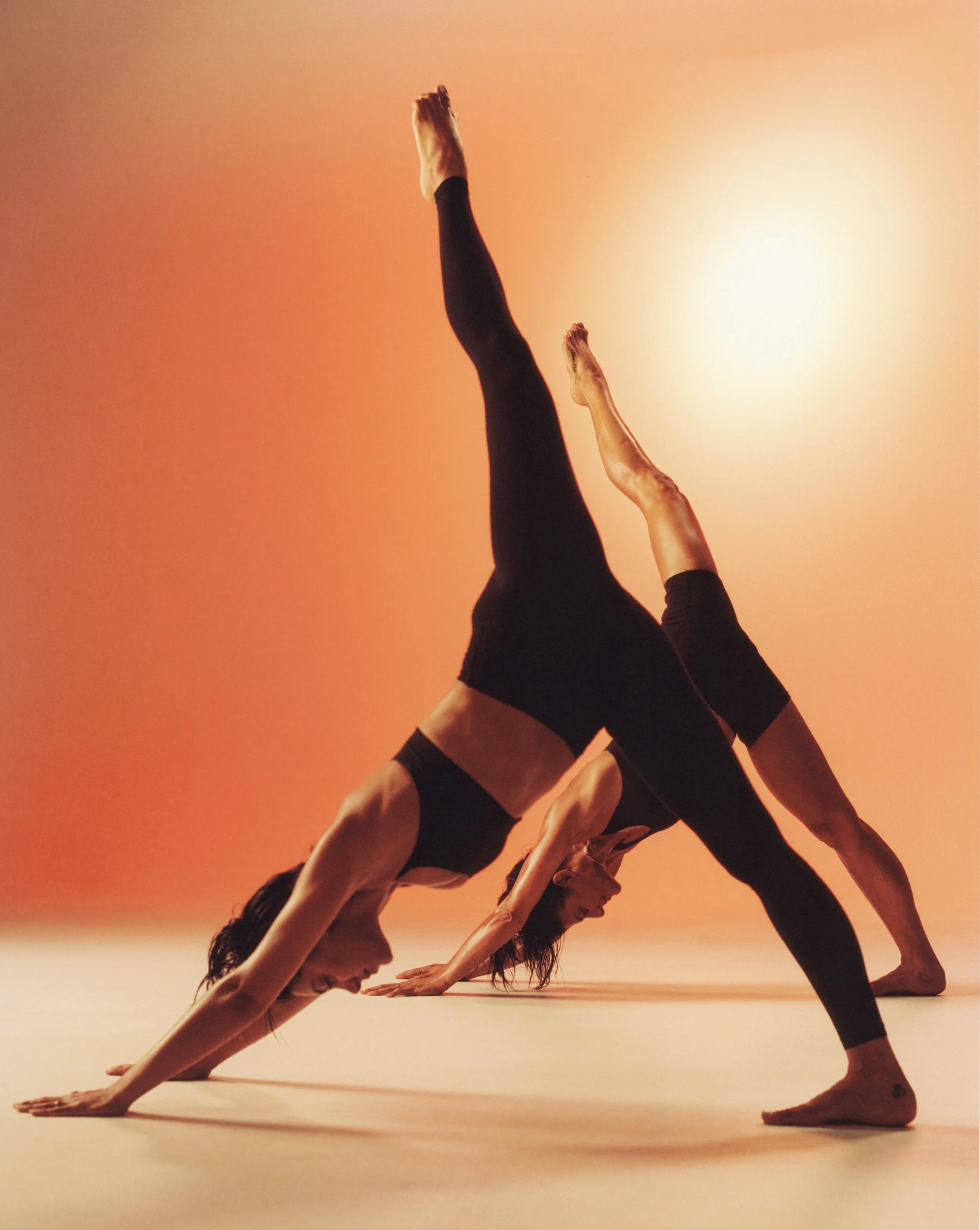 Two women practicing yoga in a downward dog position, with one woman's leg extended upward and the other woman's body parallel to the ground, against a warm, gradient background.