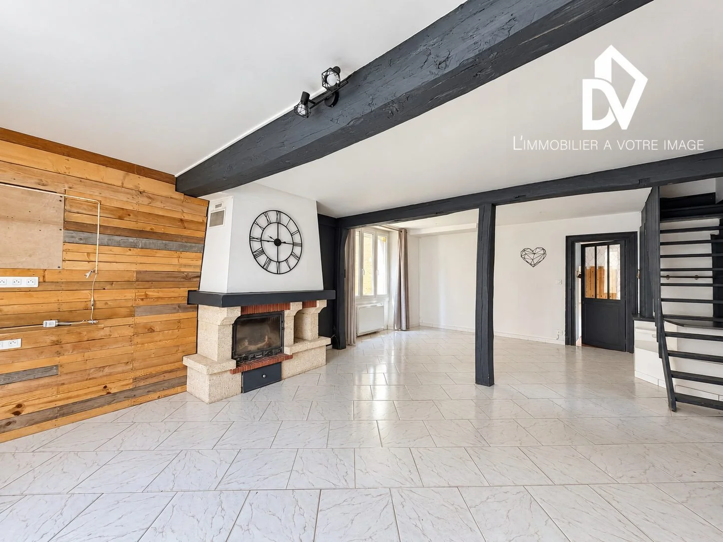 Salon lumineux avec cheminée en pierre, murs en bois, poutres apparentes noires et escalier contemporain