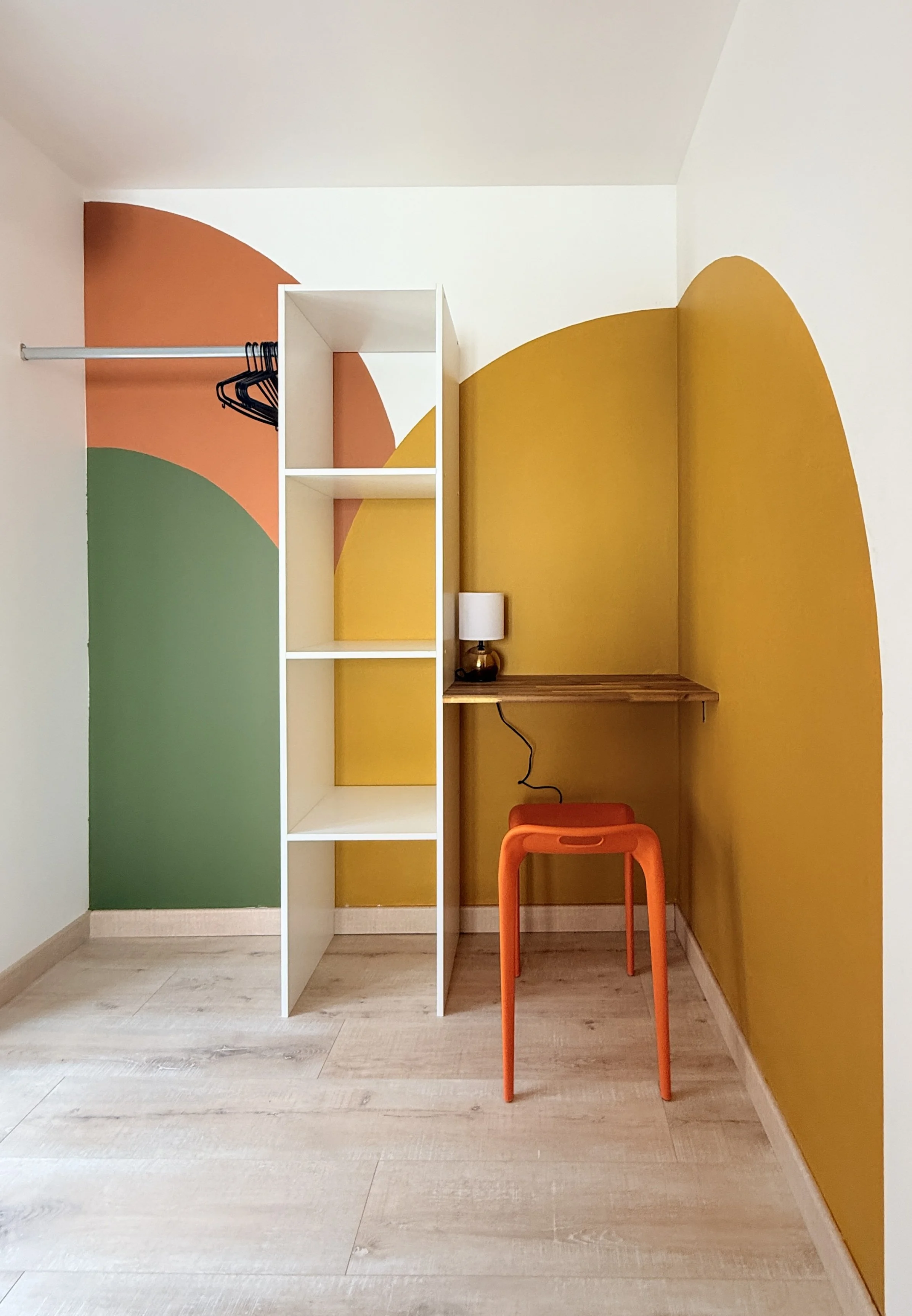 Une pièce avec un mur peint en différentes couleurs géométriques. Il y a une étagère blanche, un bureau en bois avec une lampe, une chaise orange et un mur orange et jaune en arrière-plan.