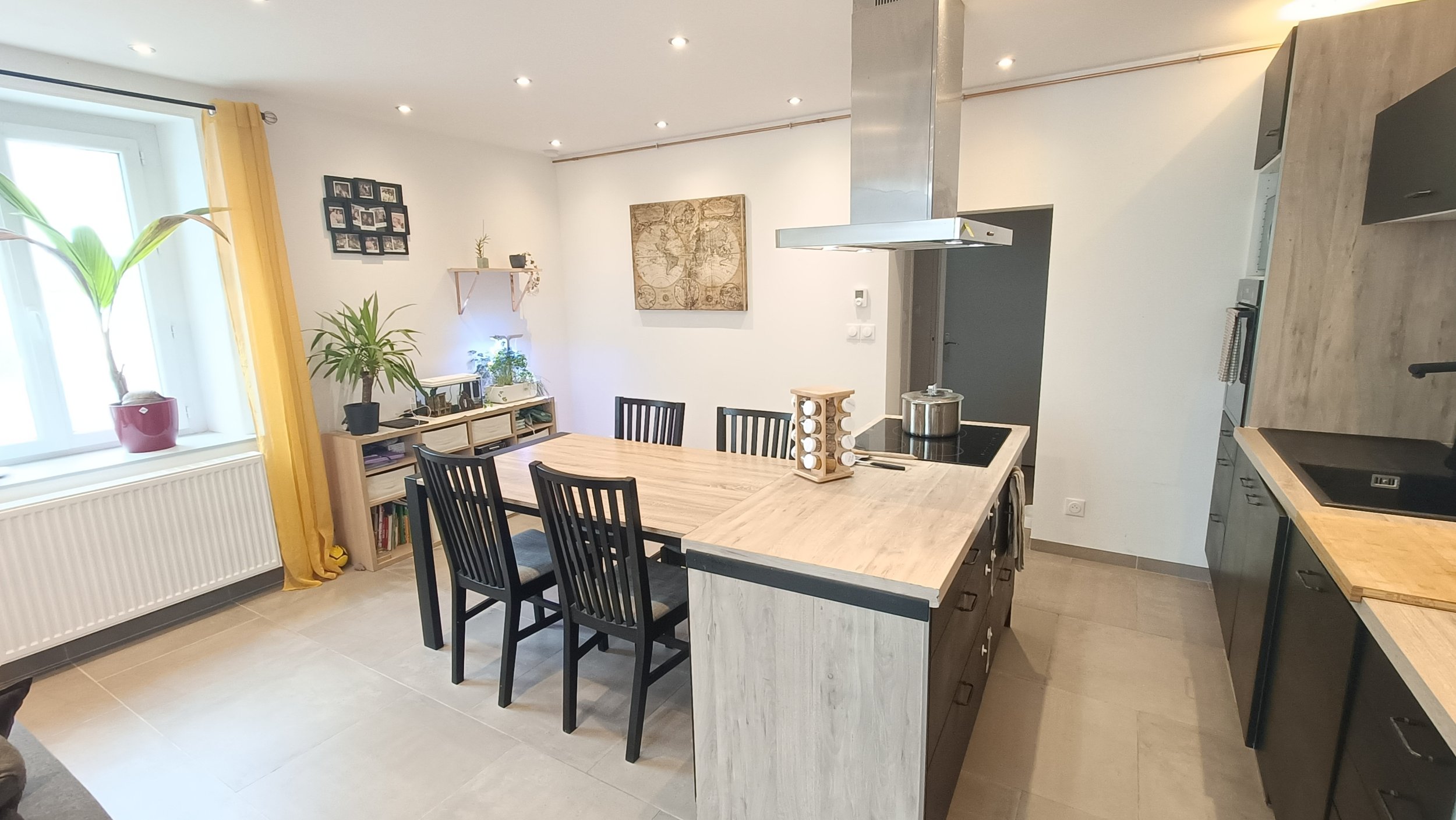 Cuisine moderne avec une table en bois, plusieurs chaises noires, un plan de travail en bois avec appareils électroménagers, et une décoration avec des plantes et des cadres