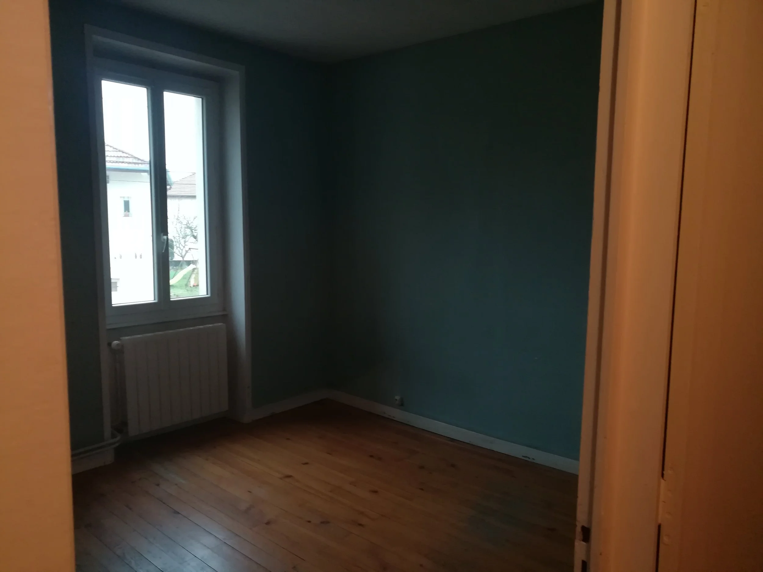 Une pièce vide avec un mur bleu, un sol en bois clair, une fenêtre laissant entrer la lumière naturelle et un radiateur sous la fenêtre.