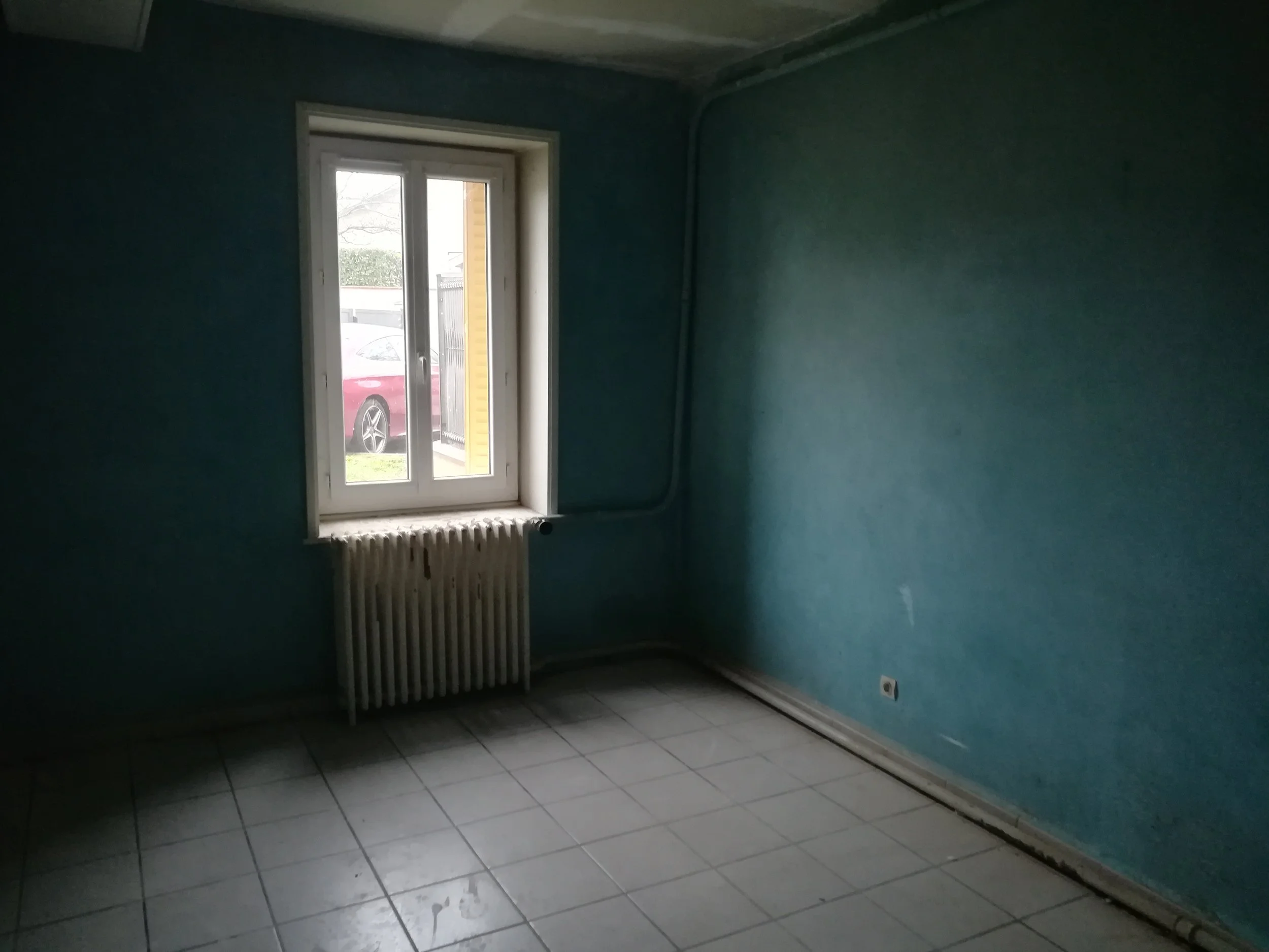 Une pièce vide avec une fenêtre et un radiateur sous la fenêtre, un mur peint en bleu vert et un sol en carreaux gris. Il y a une prise électrique à droite et une bobine de fil électrique suspendue au mur.