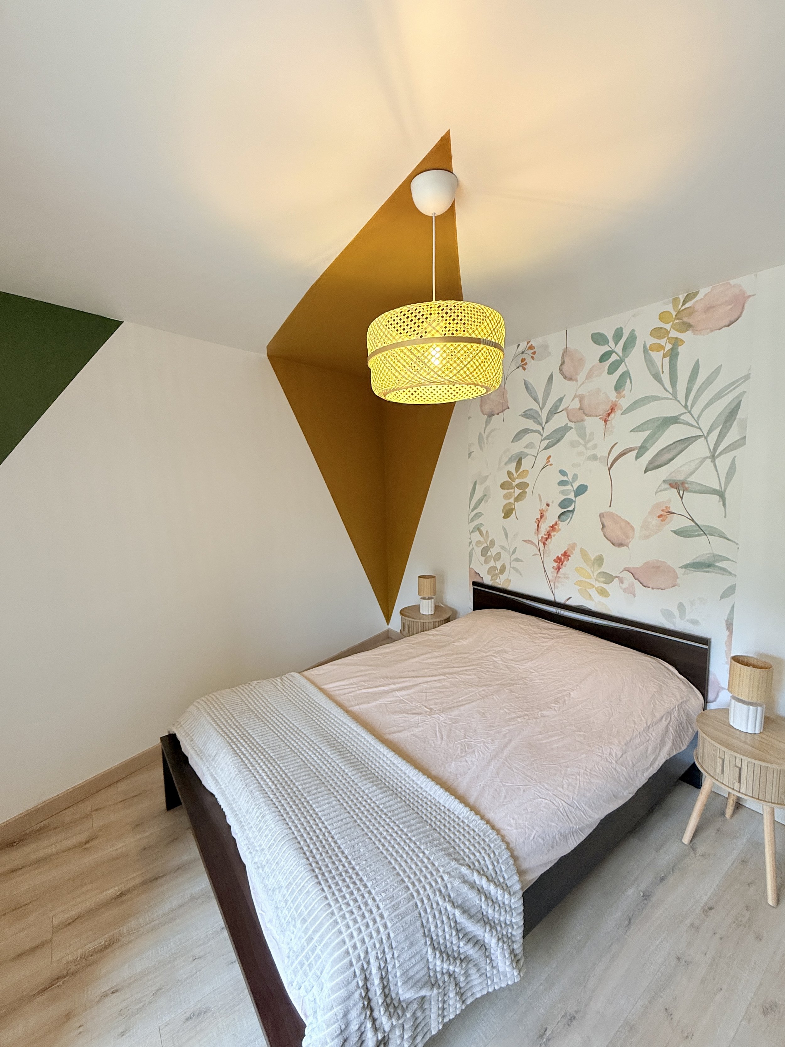 Chambre avec un lit, deux tables de chevet en bois clair, une lampe en bois, une lampe suspendue jaune au plafond et un mur avec un papier peint floral.