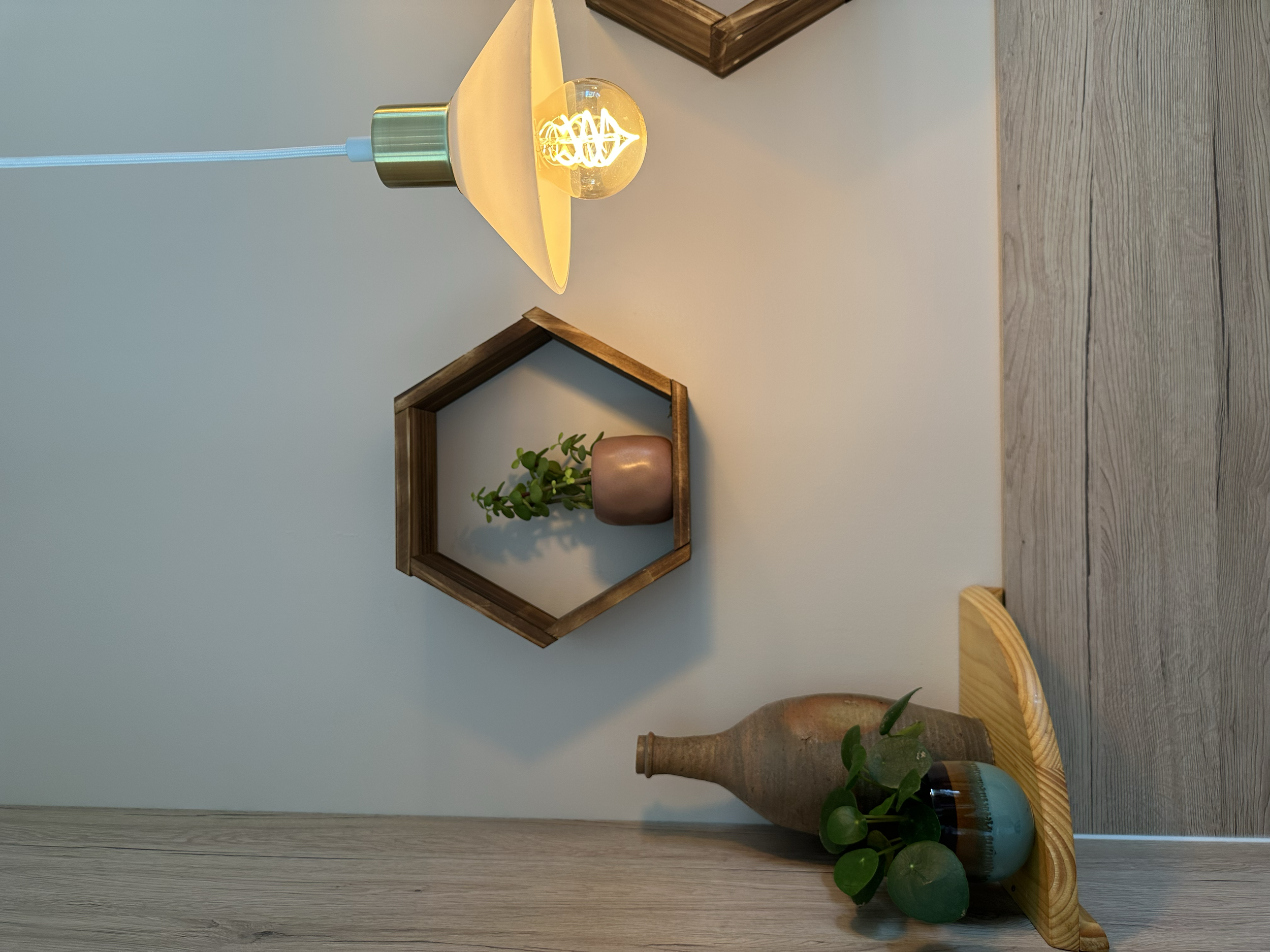 Lampe suspendue avec une ampoule à filament visible, fixée à un plafond blanc. Décorations murales avec une étagère en bois, une plante verte dans un pot et un vase beige sur un meuble en bois clair.