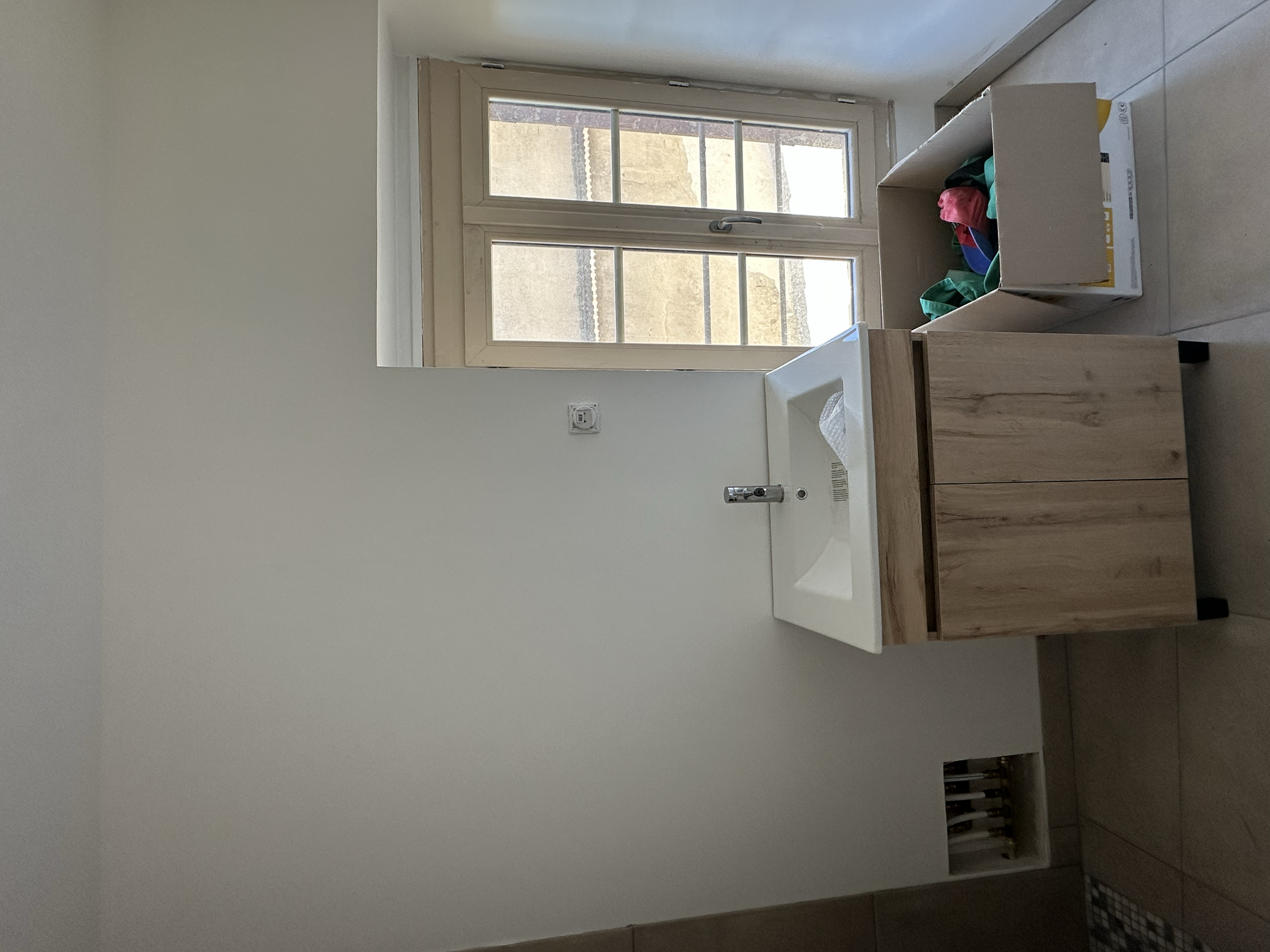 Fenêtre en bois avec plusieurs panneaux vitrés, laissant entrer la lumière dans une pièce. À côté, un lavabo encastré dans un meuble en bois. Au sol, chaussettes et autres vêtements en vrac. Mur peint en blanc, prise électrique visible.