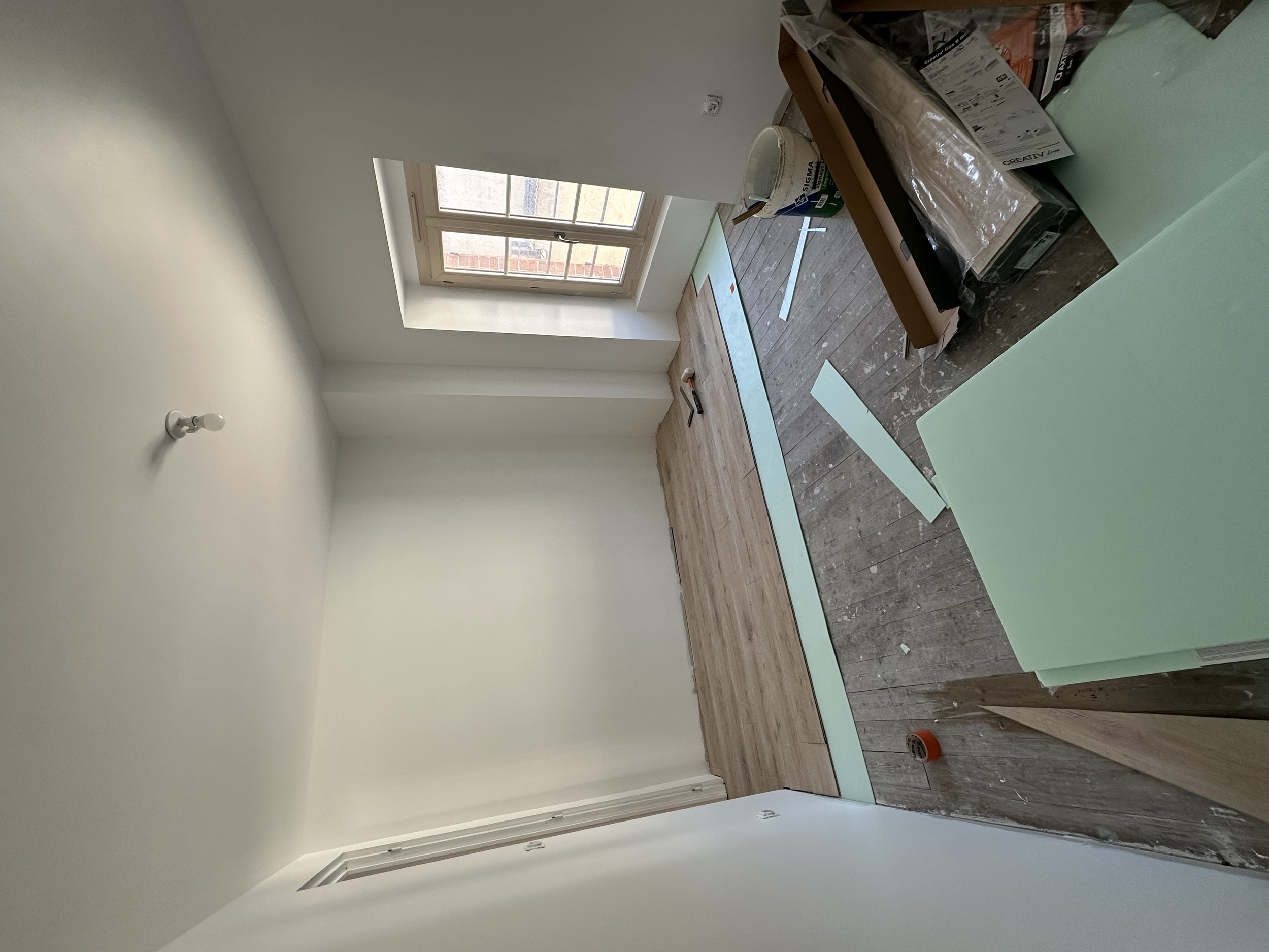 Une pièce en cours de rénovation avec un sol en bois, une fenêtre laissant entrer de la lumière, des outils et matériaux de construction sur une table, et du papier de protection vert.
