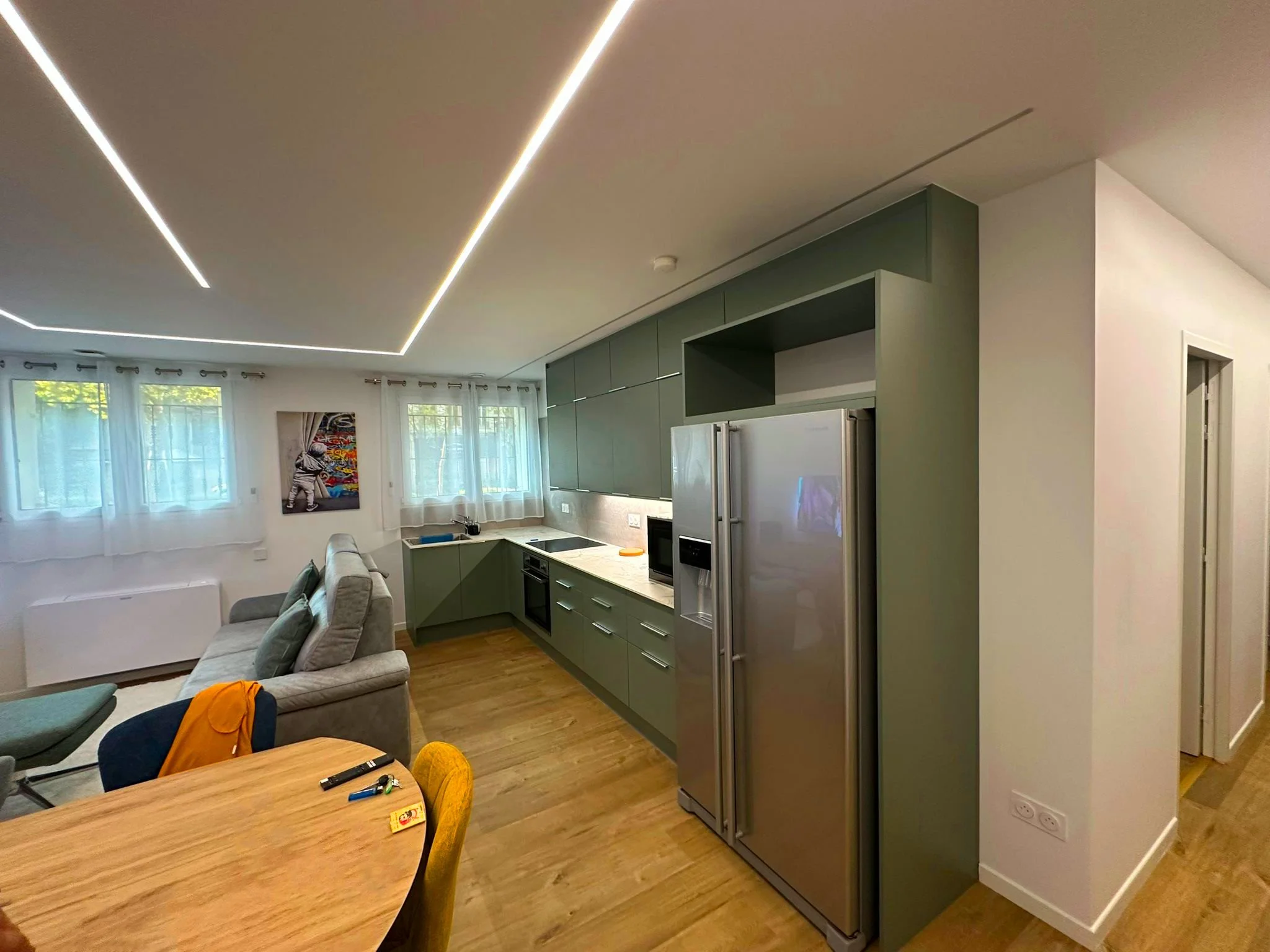 Intérieur d'une cuisine moderne avec murs verts, armoires vertes, grand réfrigérateur en acier inoxydable, zone de cuisson, et salon avec canapé gris près de fenêtres avec rideaux blancs graphiques.