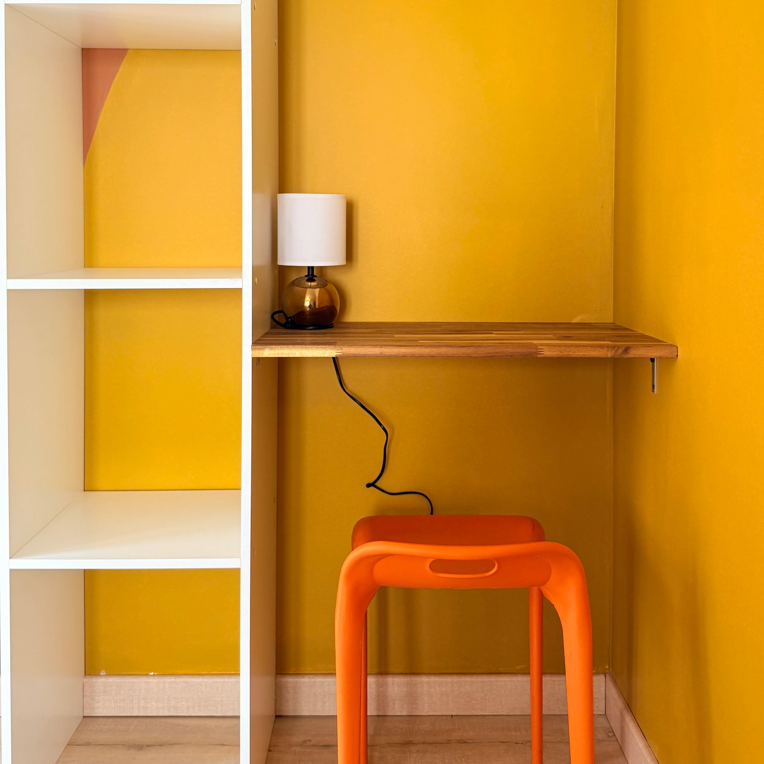 Une étagère blanche, un tableau jaune, une lampe blanche sur une table en bois, une chaise orange, un mur jaune.
