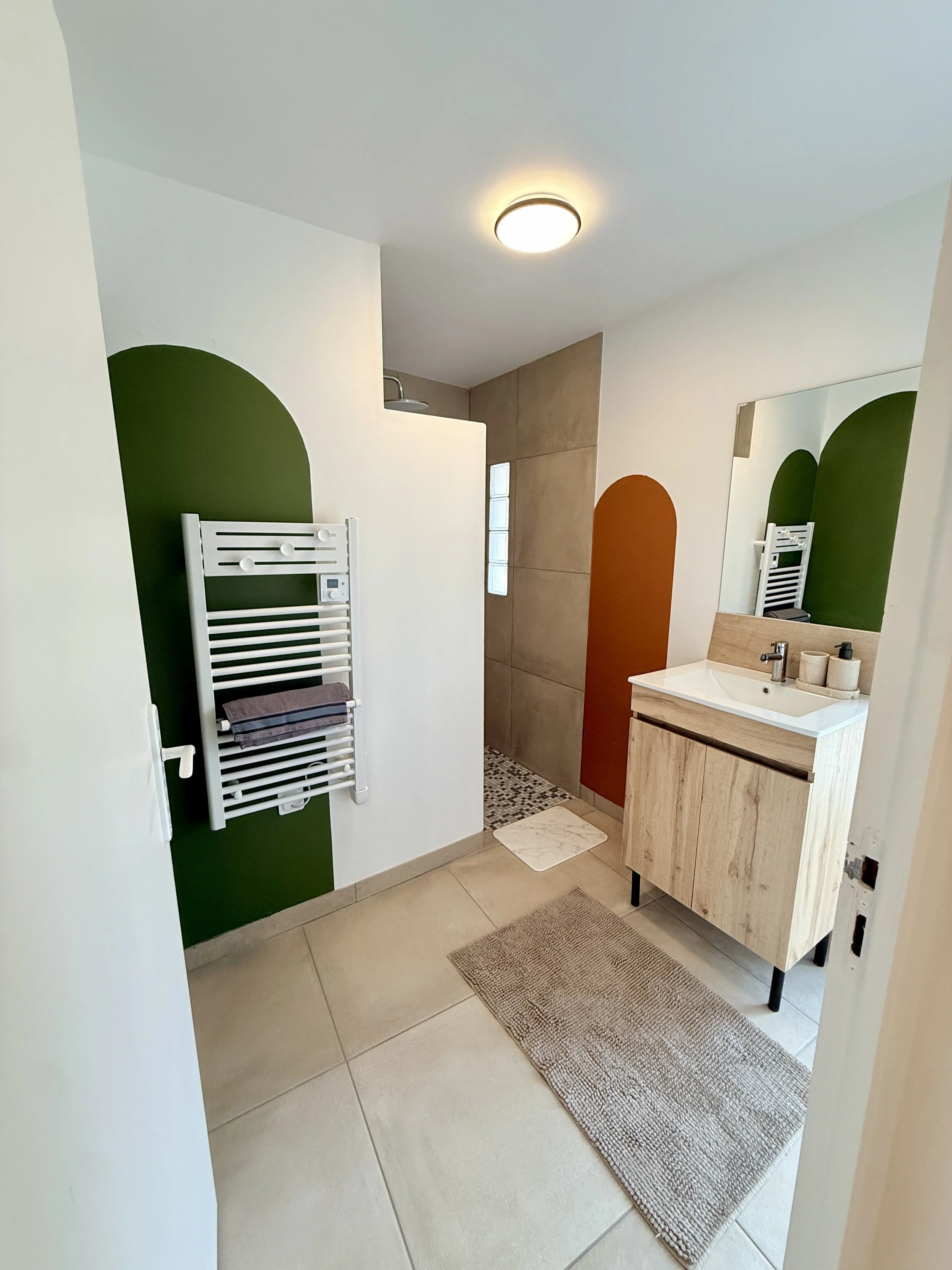 Salle de bain moderne avec un meuble en bois, un lavabo blanc, un miroir, des tapis, un radiateur parking ainsi qu'une douche dans un coin.