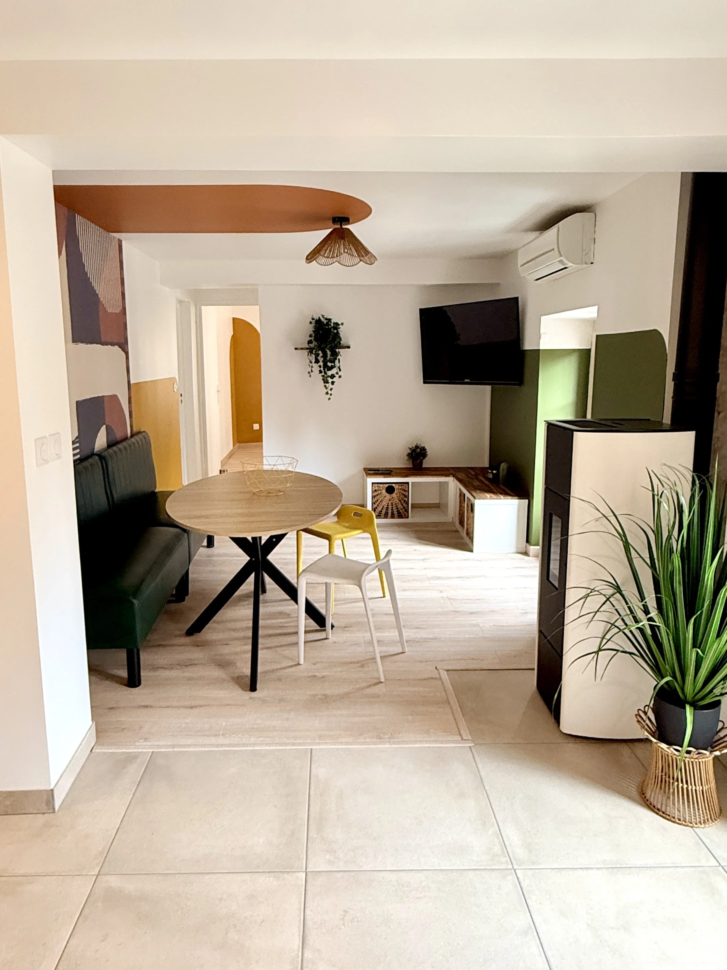 Salon avec canapé vert, table ronde, chaises jaunes et blanches, télévision murale, étagère avec plantes, grand vase avec plante, mur blanc, plafond avec lampe en forme de fleur, sol en bois.