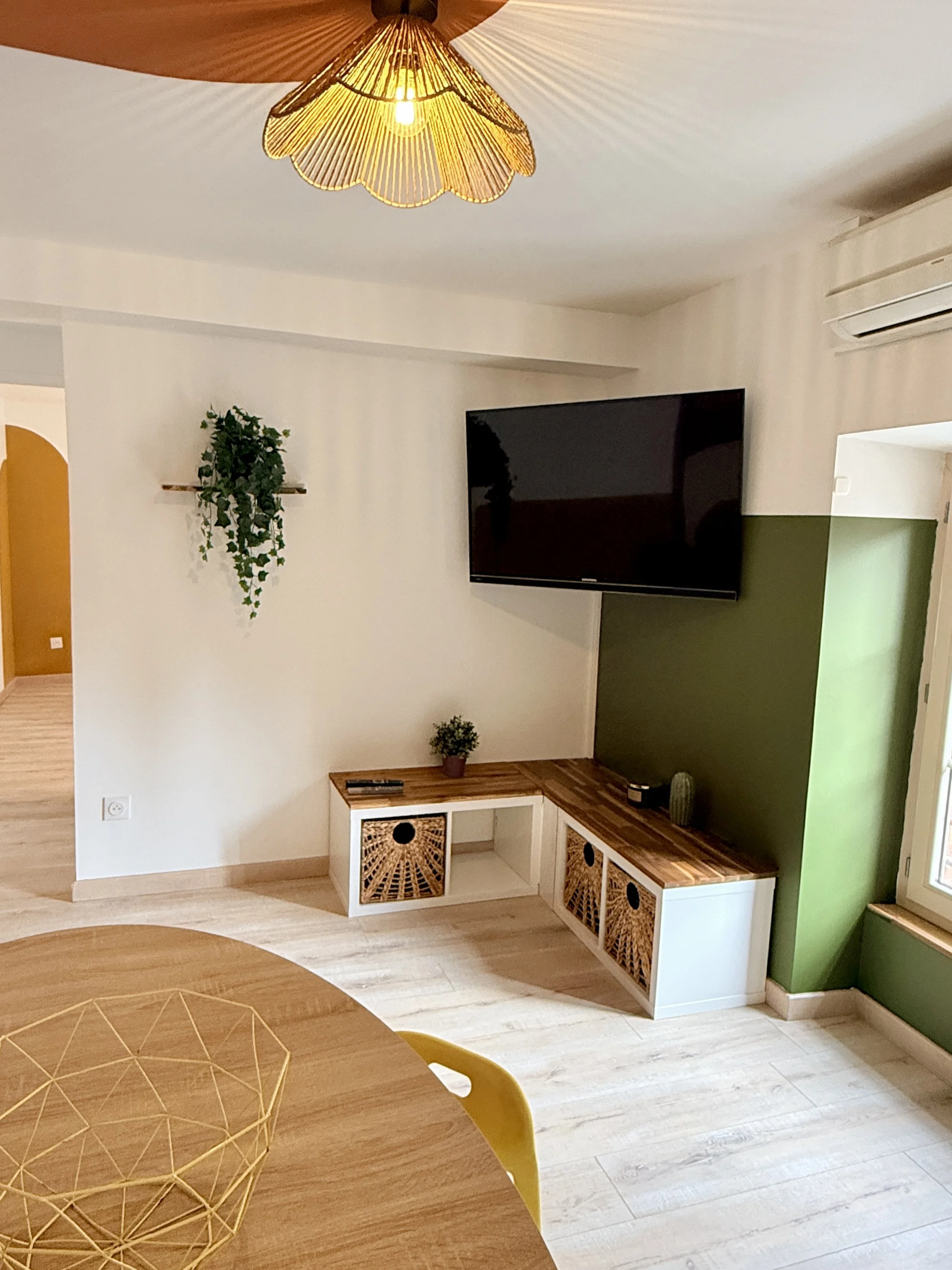 Un salon moderne avec un mur blanc, une télévision à écran plat, un meuble en bois avec des paniers en rotin, une plante suspendue, et un éclairage chaud suspendu au plafond.