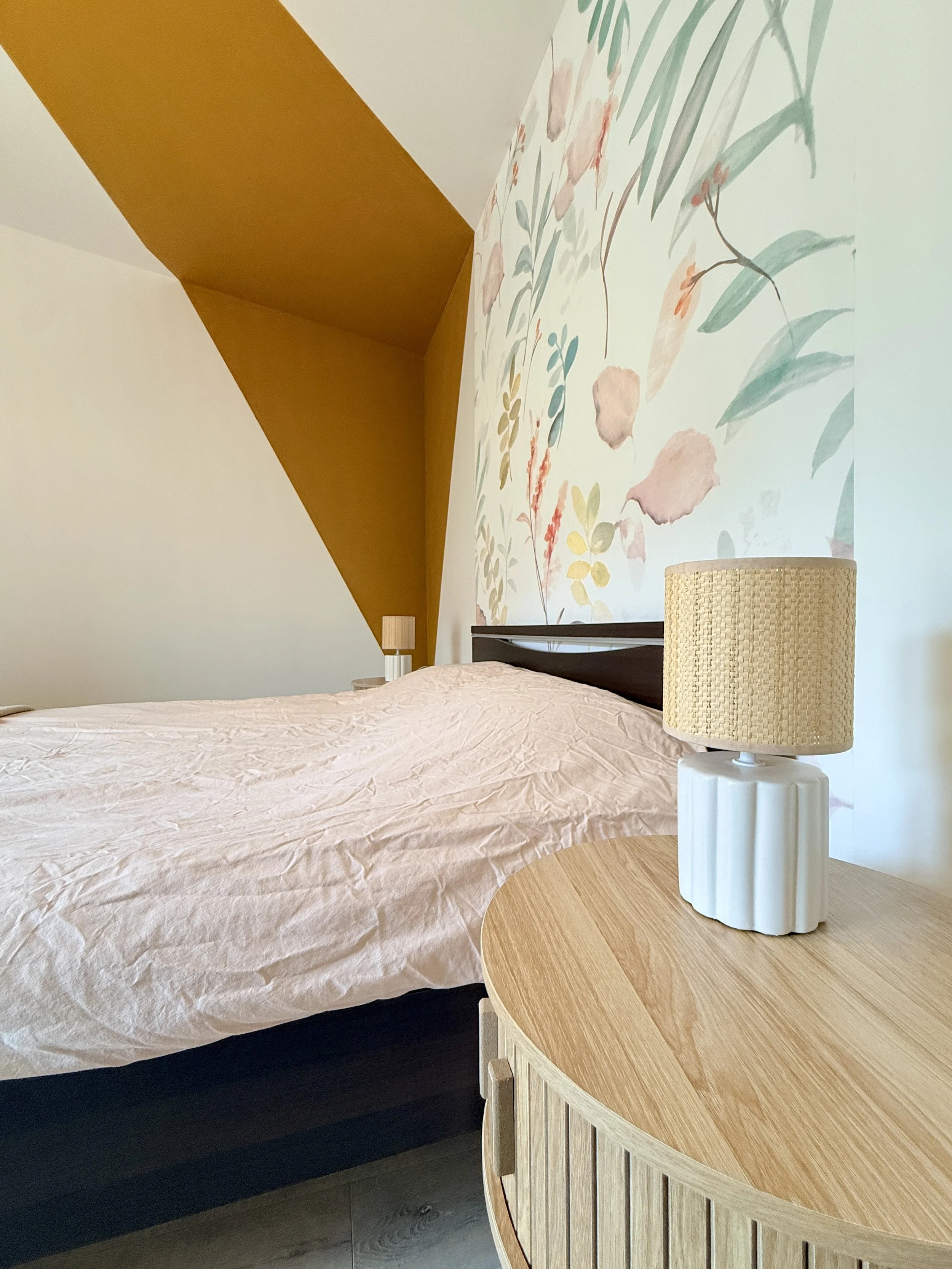 Une chambre avec un lit, une table de chevet avec une lampe, une tête de lit et un mur avec un motif floral pastel.