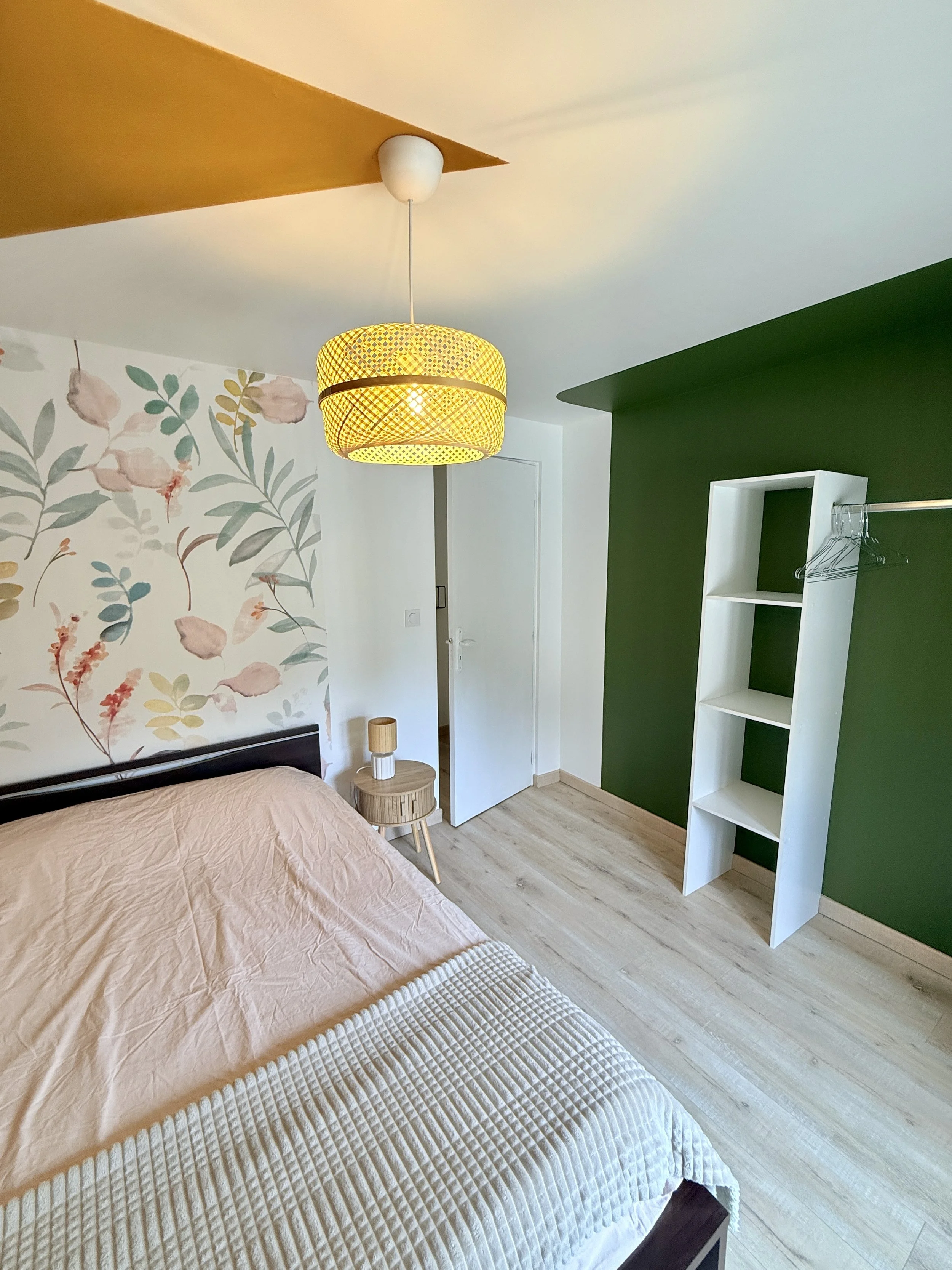 Chambre avec un lit, un mur avec un papier peint floral, une suspension lumineuse jaune, une petite table de chevet, une étagère blanche et un mur vert.