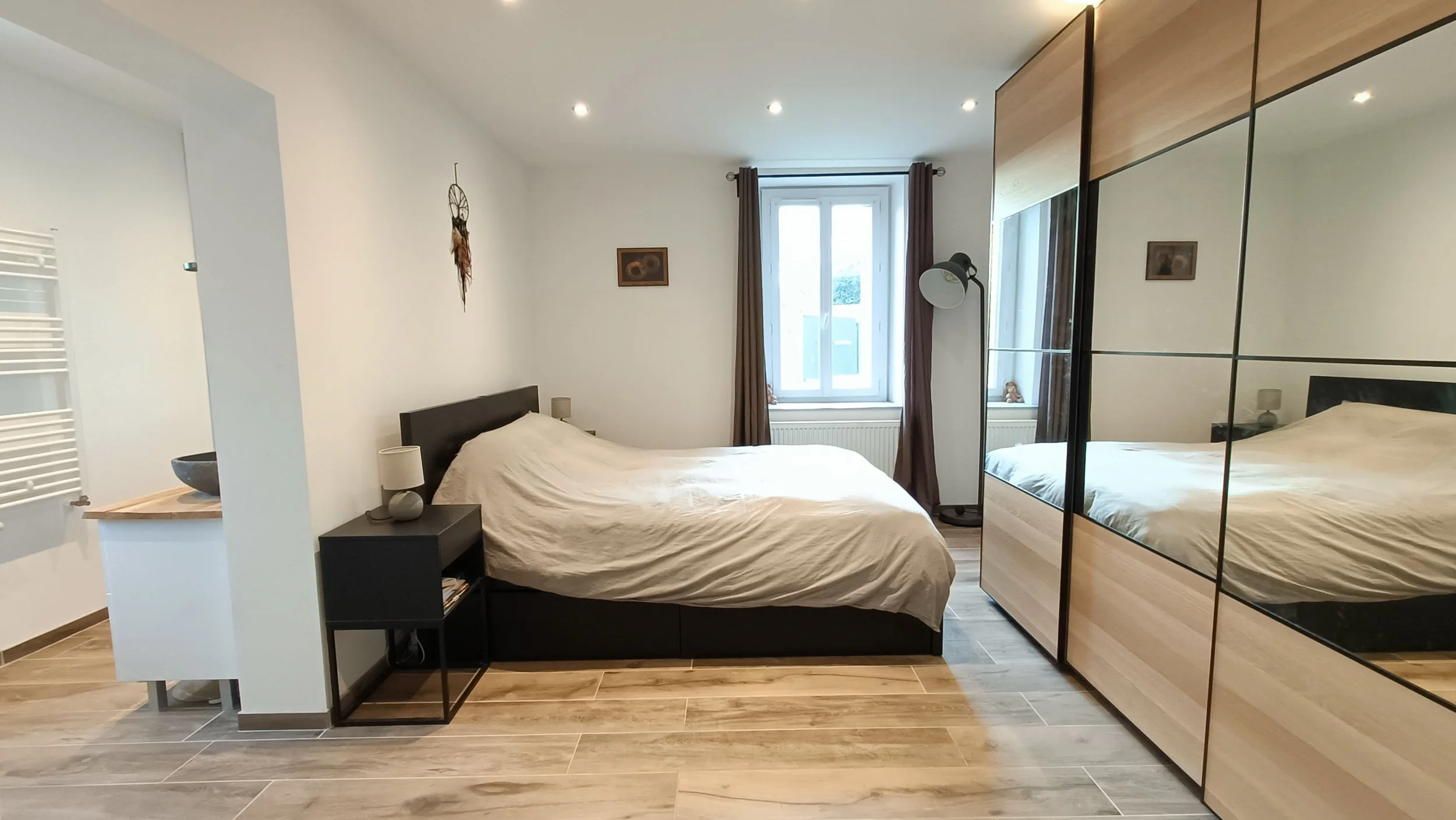 Chambre moderne avec un lit double, un grand placard en miroir, une fenêtre avec rideaux, une lampe de lecture, et des décorations murales.