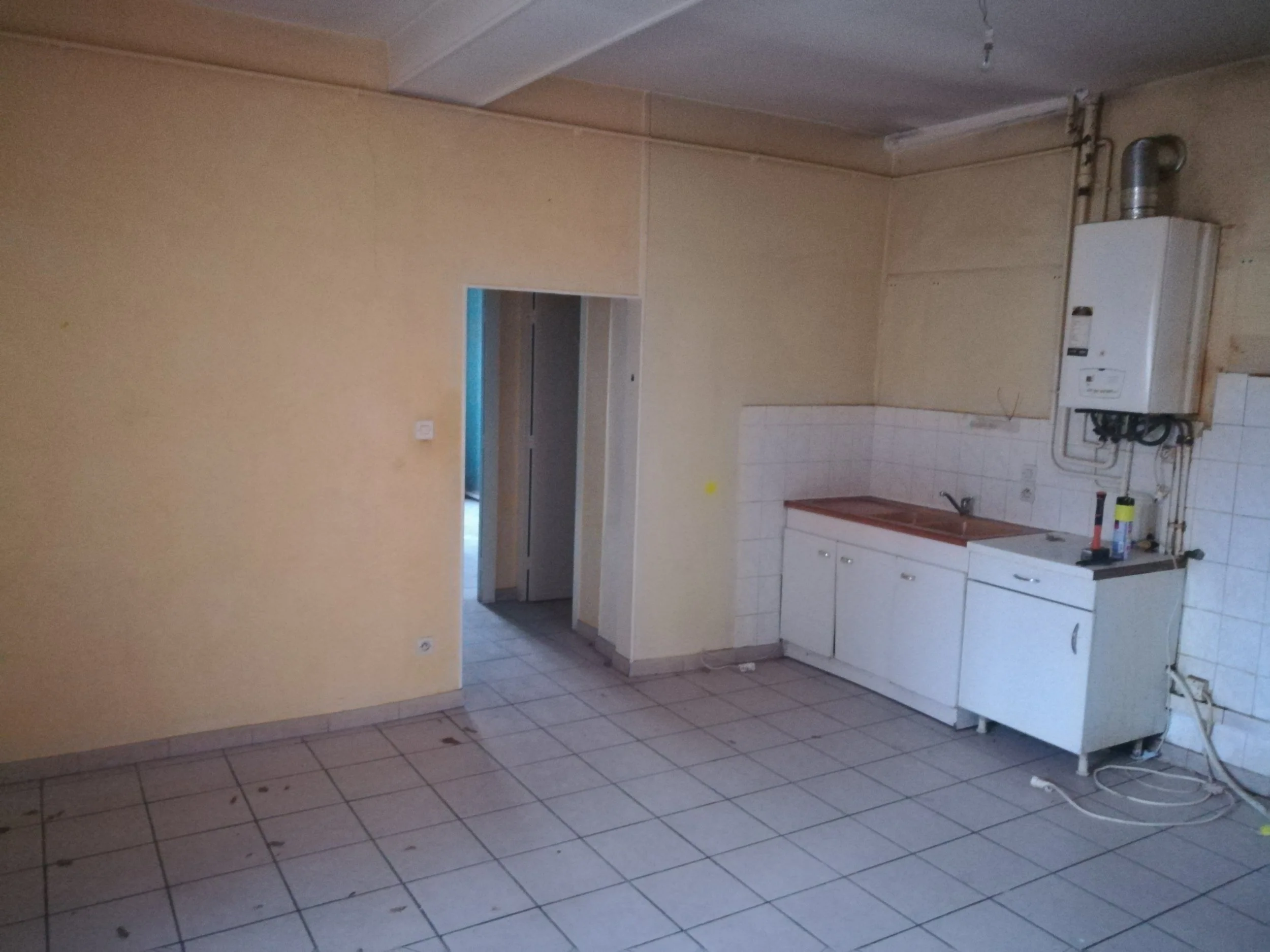 Une pièce vide avec un mur beige, un carrelage gris au sol, et une cuisine blanche avec des meubles bas. Un chauffe-eau est fixé au mur. Il y a un passage vers une autre pièce ou un couloir à gauche.