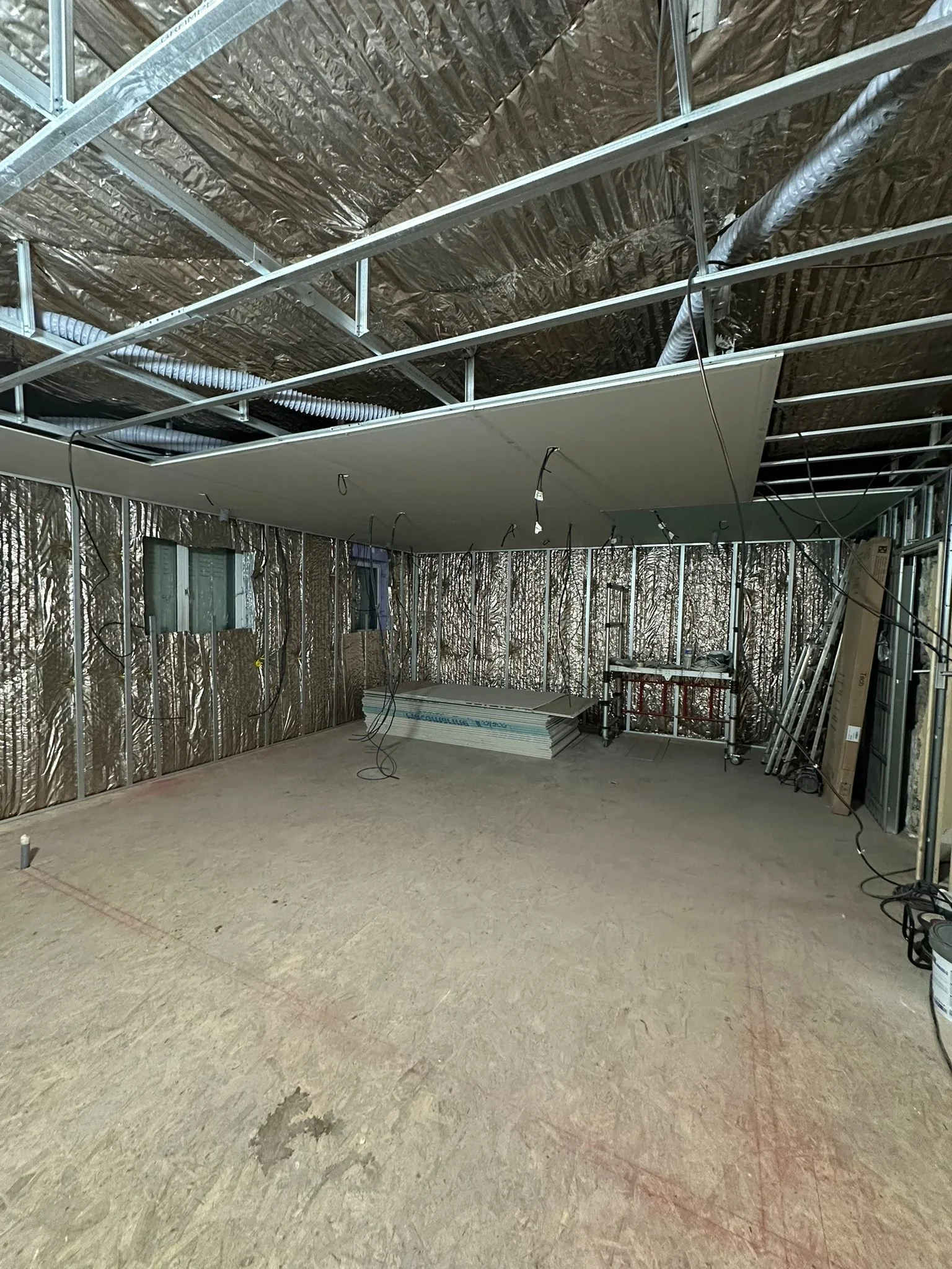 Intérieur d'un bâtiment en construction avec murs isolés en aluminium, plafond suspendu en cours d'installation, câbles électriques pendants, sol en stratifié, osb empilé, fenêtres et échelles visibles.