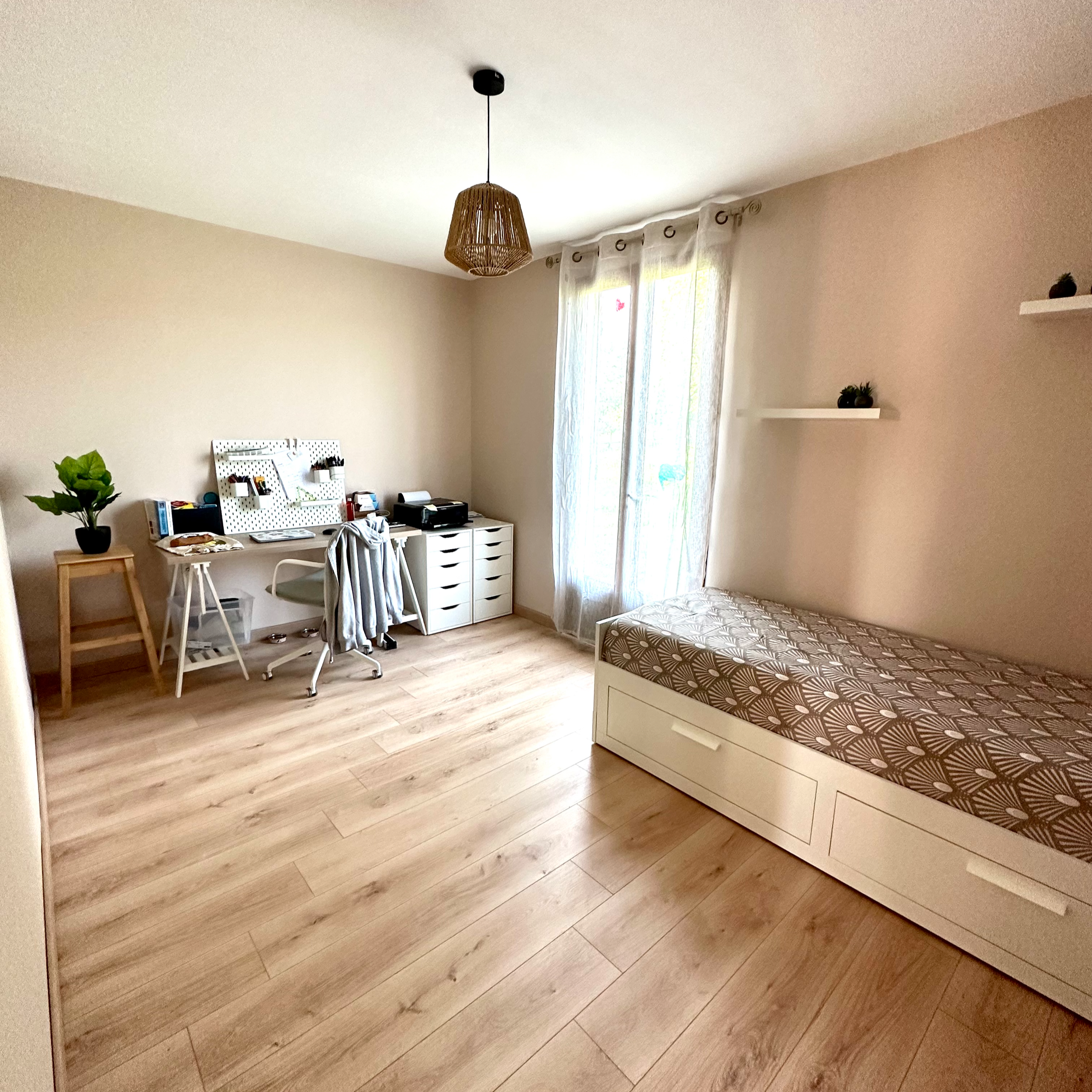 Une chambre avec un lit à cadre blanc, un bureau, une plante en pot, des étagères blanches avec des objets decoratifs et une fenêtre avec des rideaux blancs, et un plafonnier en rotin.