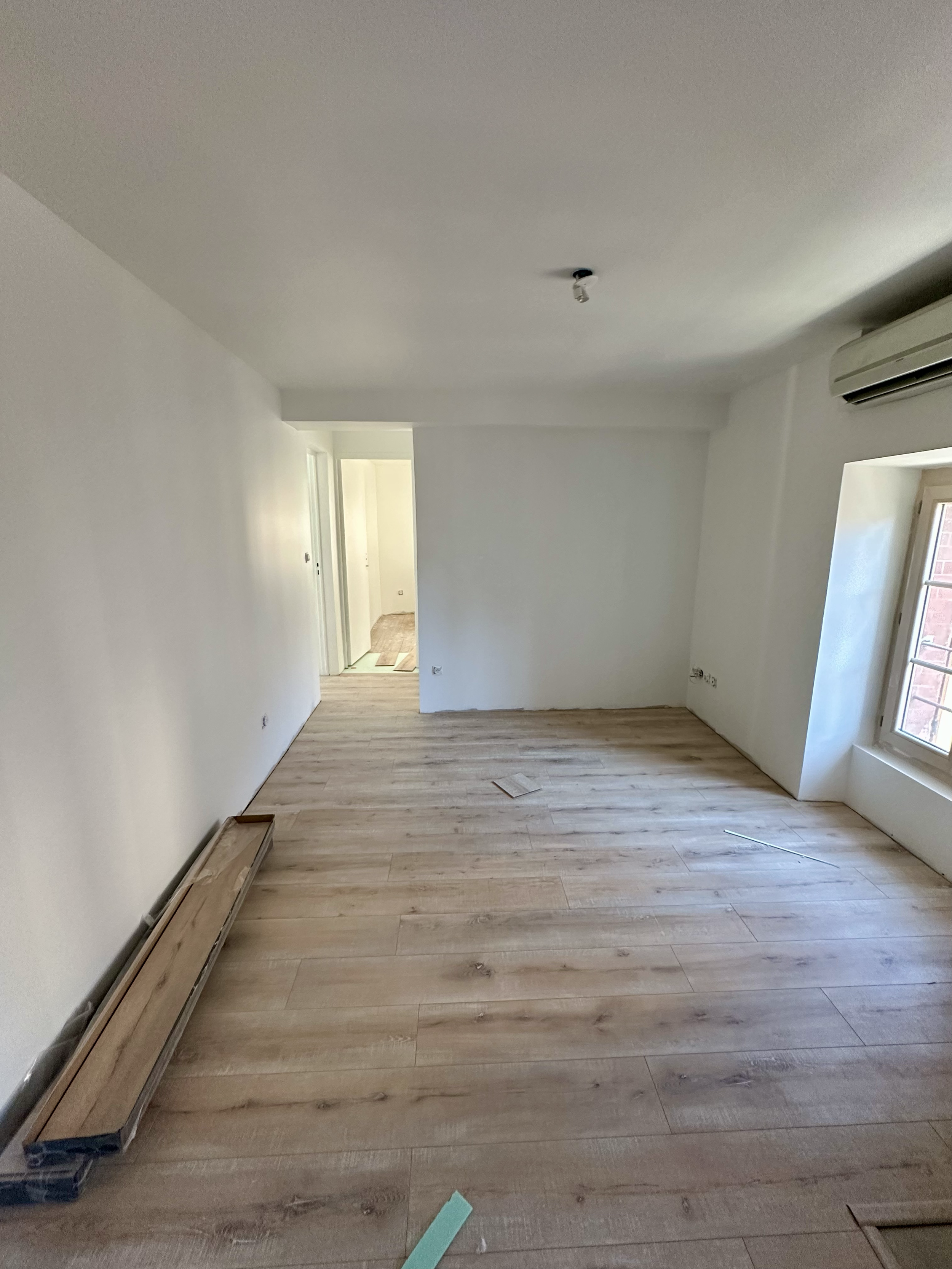 Une pièce en cours de rénovation avec un sol en bois clair, des murs blanc, une fenêtre avec volet en bois, un climatiseur et un plafond sans luminaire, avec des éléments de construction au sol.