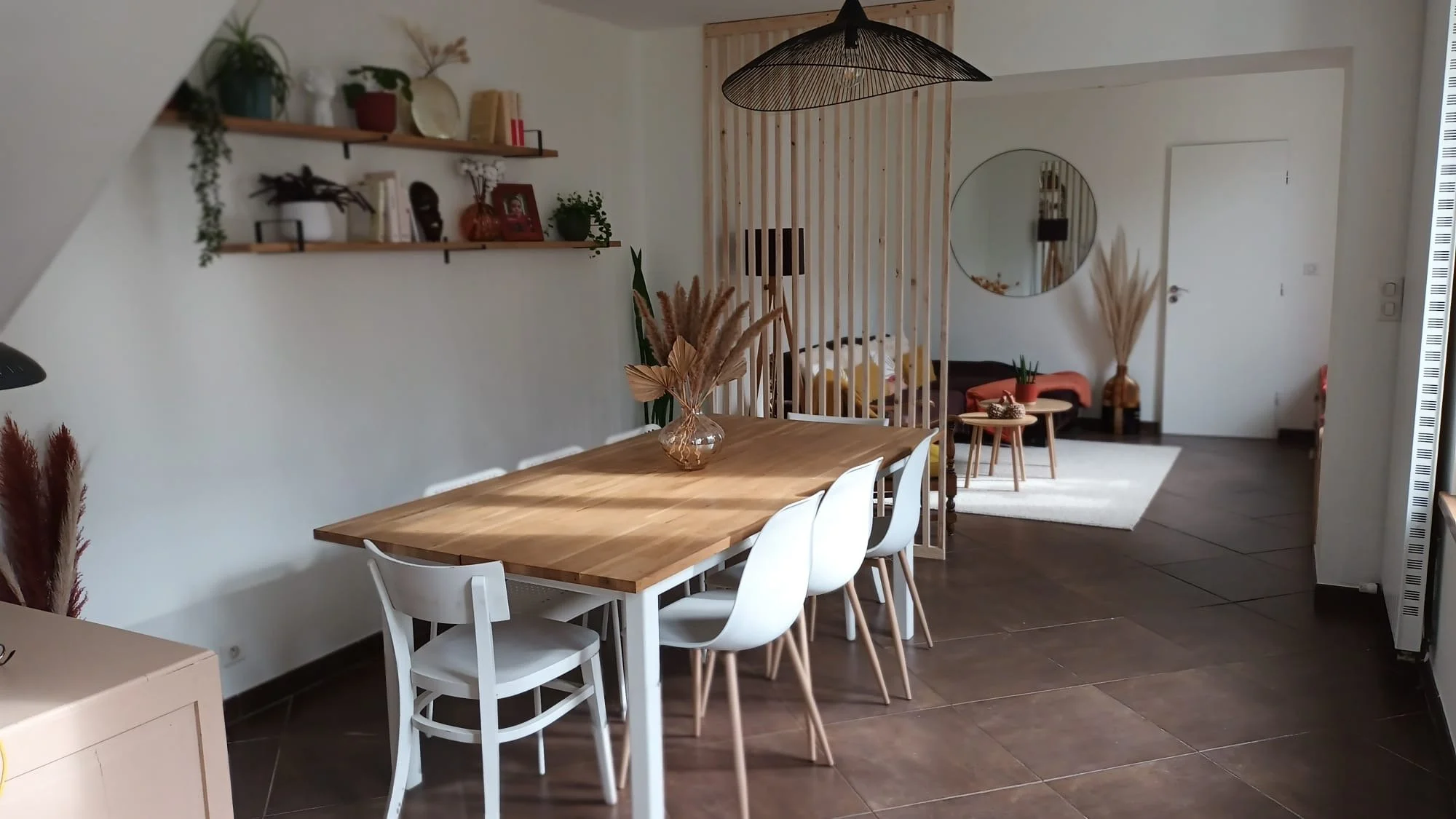 Intérieur d'un salon avec une table en bois entourée de chaises blanches, décorations végétales, étagères avec des objets décoratifs, un canapé avec des coussins jaunes, un grand miroir rond, et un lambris en bois séparant différentes zones du salon.