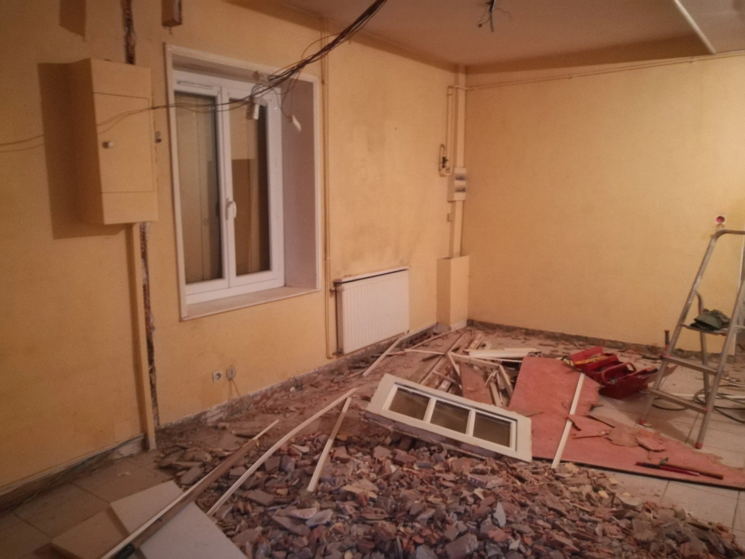 Une pièce en travaux avec des débris de plâtre et un cadre de fenêtre cassé sur le sol, murs jaunes et une porte-fenêtre.