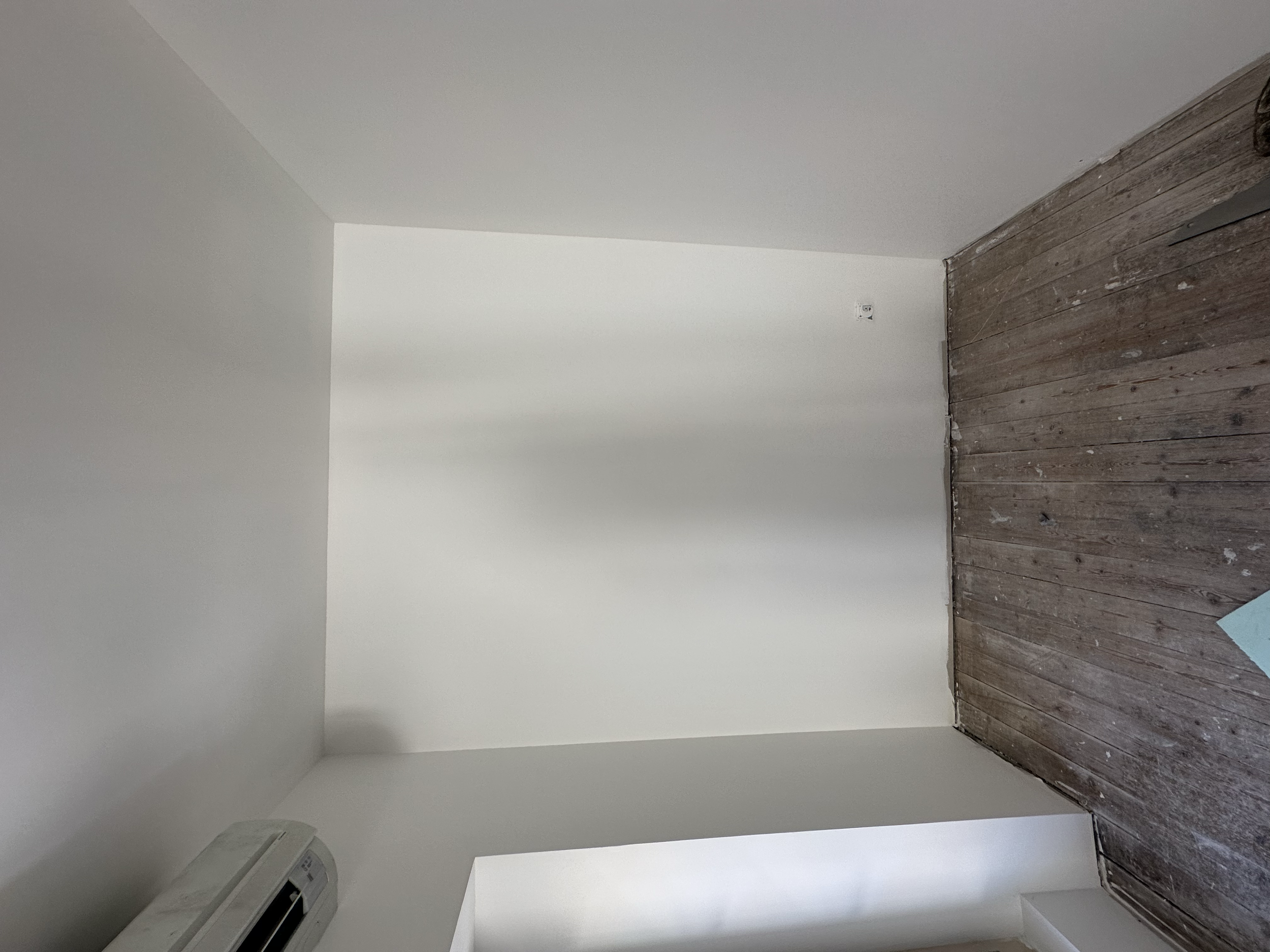 Une pièce avec murs blancs, un plancher en bois ancien, et une unité de climatisation murale dans le coin inférieur gauche.