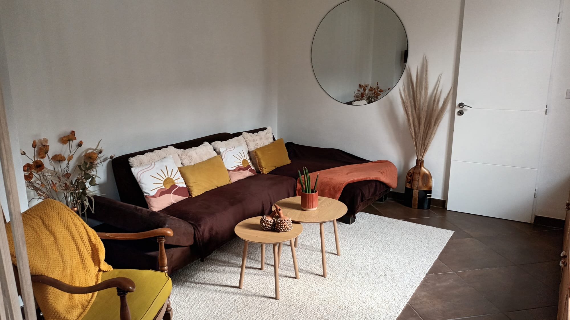 Salon avec un canapé marron, coussins à motifs de soleil, une petite table en bois avec un vase à plantes, un fauteuil en bois avec un coussin jaune, une grande vase contenant des pampas, miroir rond au mur, porte blanche, tapis blanc, décoration mur