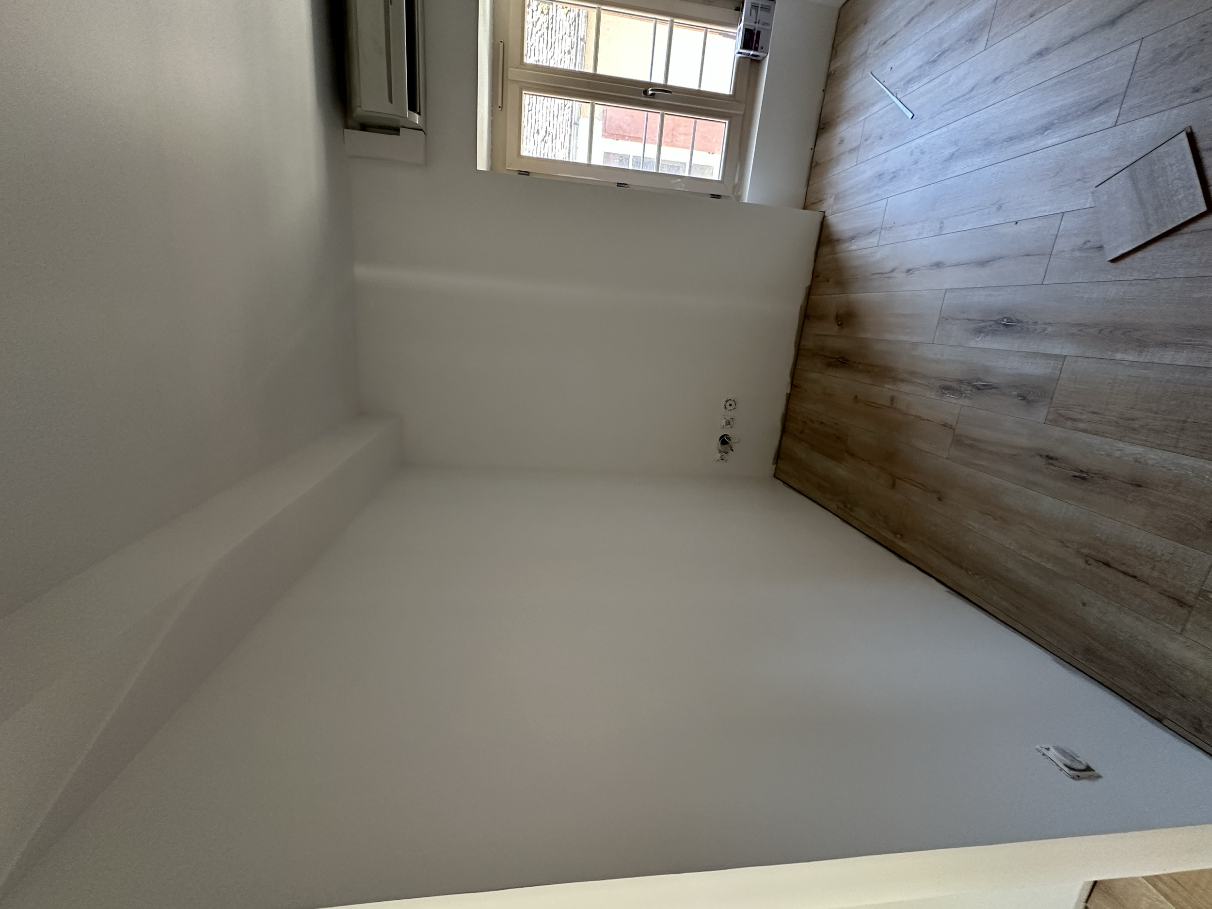 Une pièce en cours de rénovation avec un parquet en bois clair, un mur blanc et une fenêtre. Le sol a des plinthes en bois. Il y a des prises électriques et un câblage visible sur le mur.