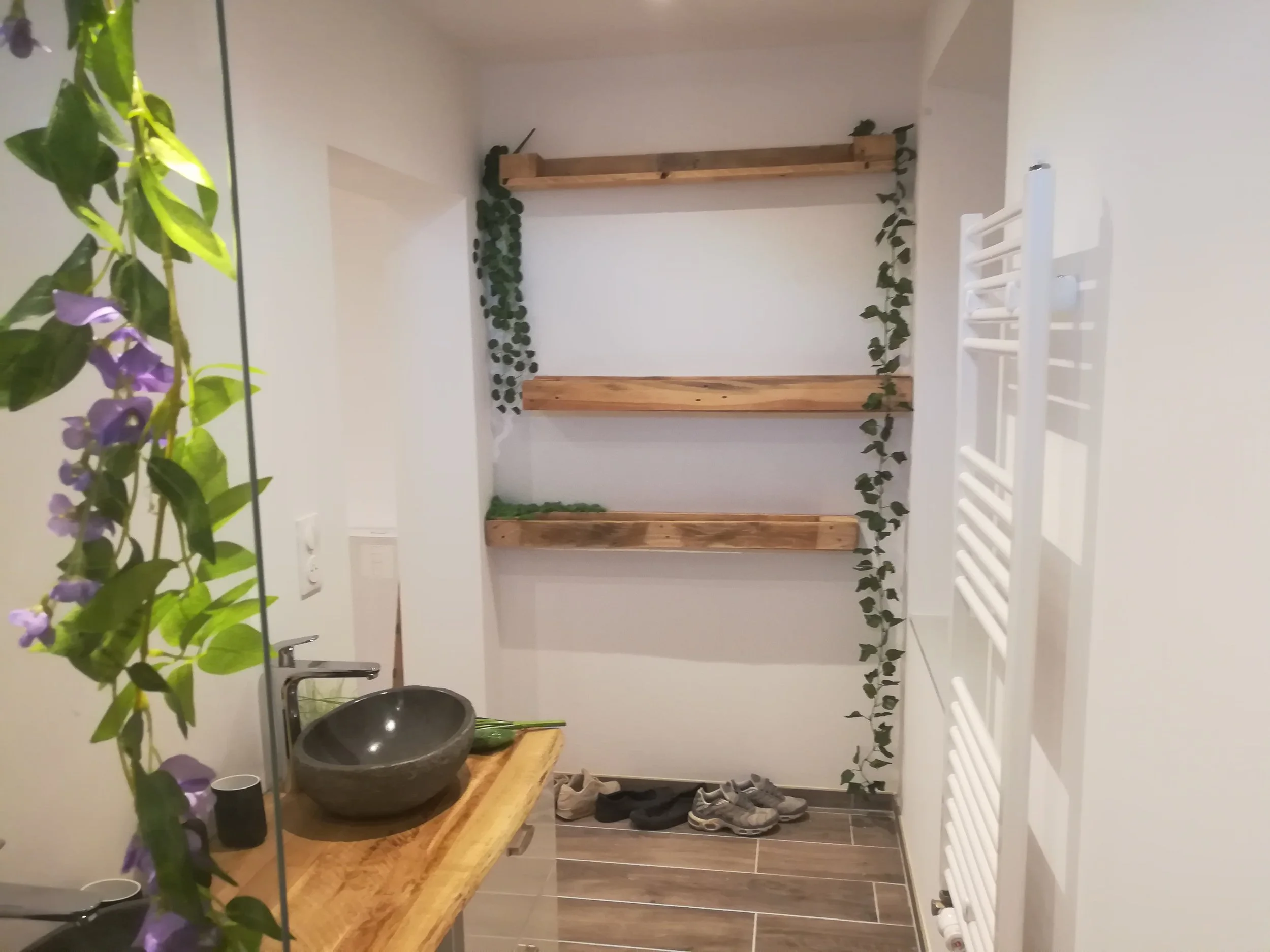 Vue intérieure d'une pièce avec étagères en bois, plantes grimpantes, un lavabo en pierre, un radiateur, des chaussures au sol et un miroir.