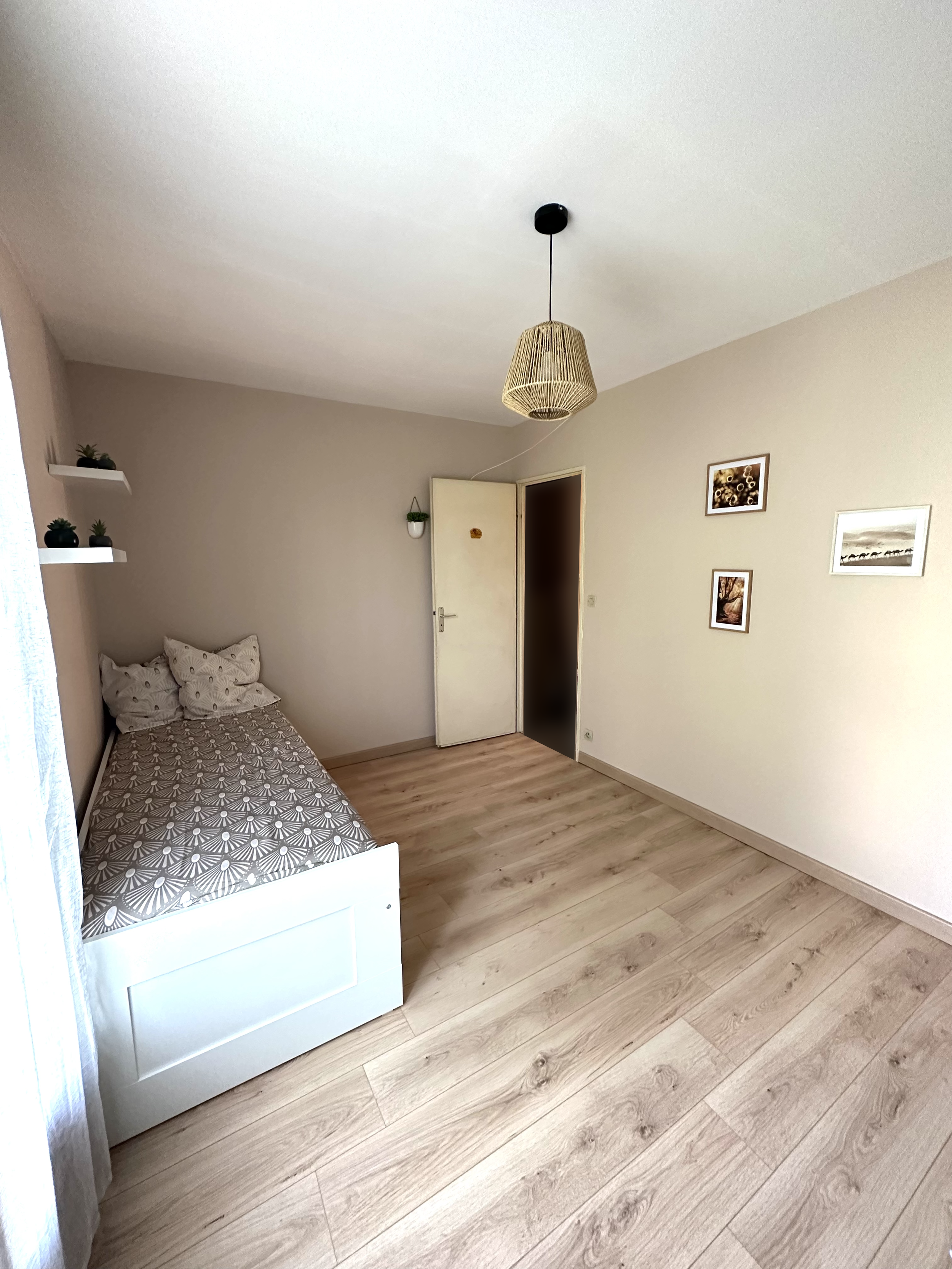 Une chambre simple avec un lit à une seule personne, des oreillers à motifs, une porte et quelques images décoratives sur le mur, un lampadaire en suspension en rotin et quelques plantes sur des étagères.