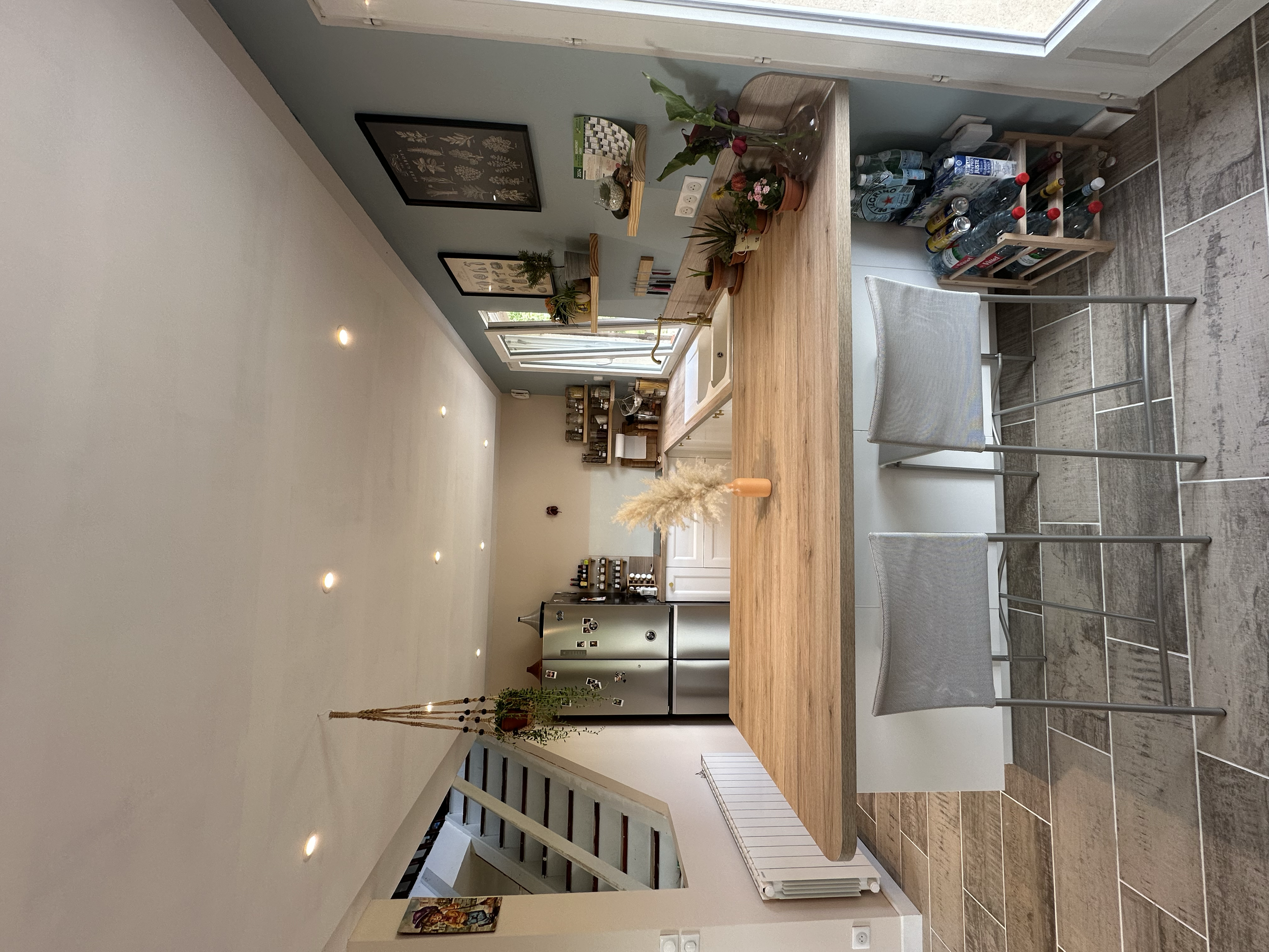 Une cuisine moderne avec un plan de travail en bois naturel, plusieurs plantes en pot, une étagère avec des livres et des accessoires, une grande fenêtre, un réfrigérateur, une chaise en métal avec un coussin, et un sol en carrelage gris.