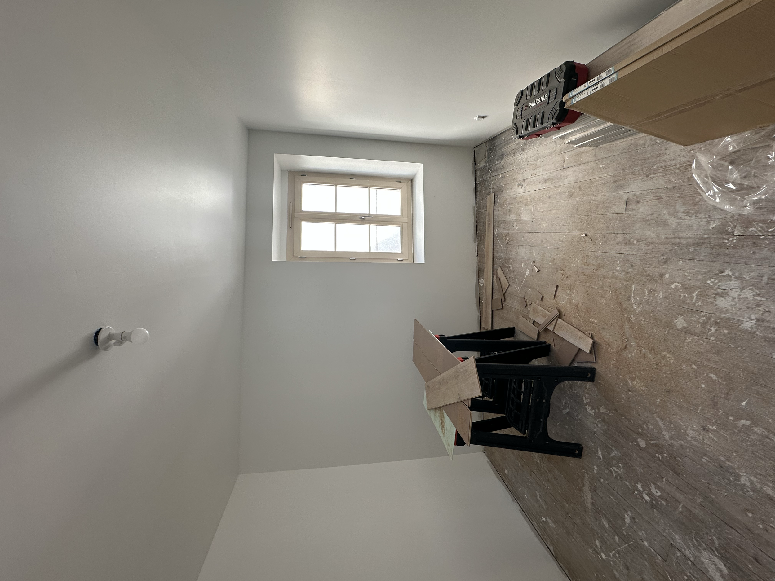 Une pièce en cours de rénovation avec un mur blanc, une fenêtre, un sol en bois, des outils et des matériaux de construction.