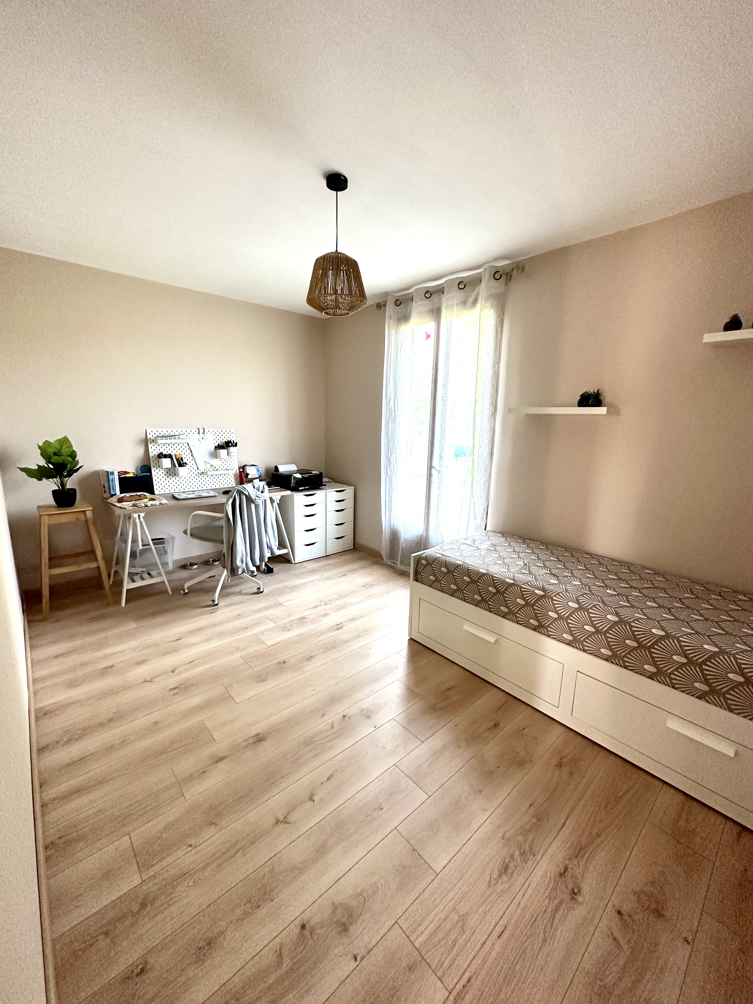 Chambre avec un lit, une étagère, une lampe en rotin suspendue, un bureau avec une plante, une chaise, et des rideaux blancs devant une fenêtre.