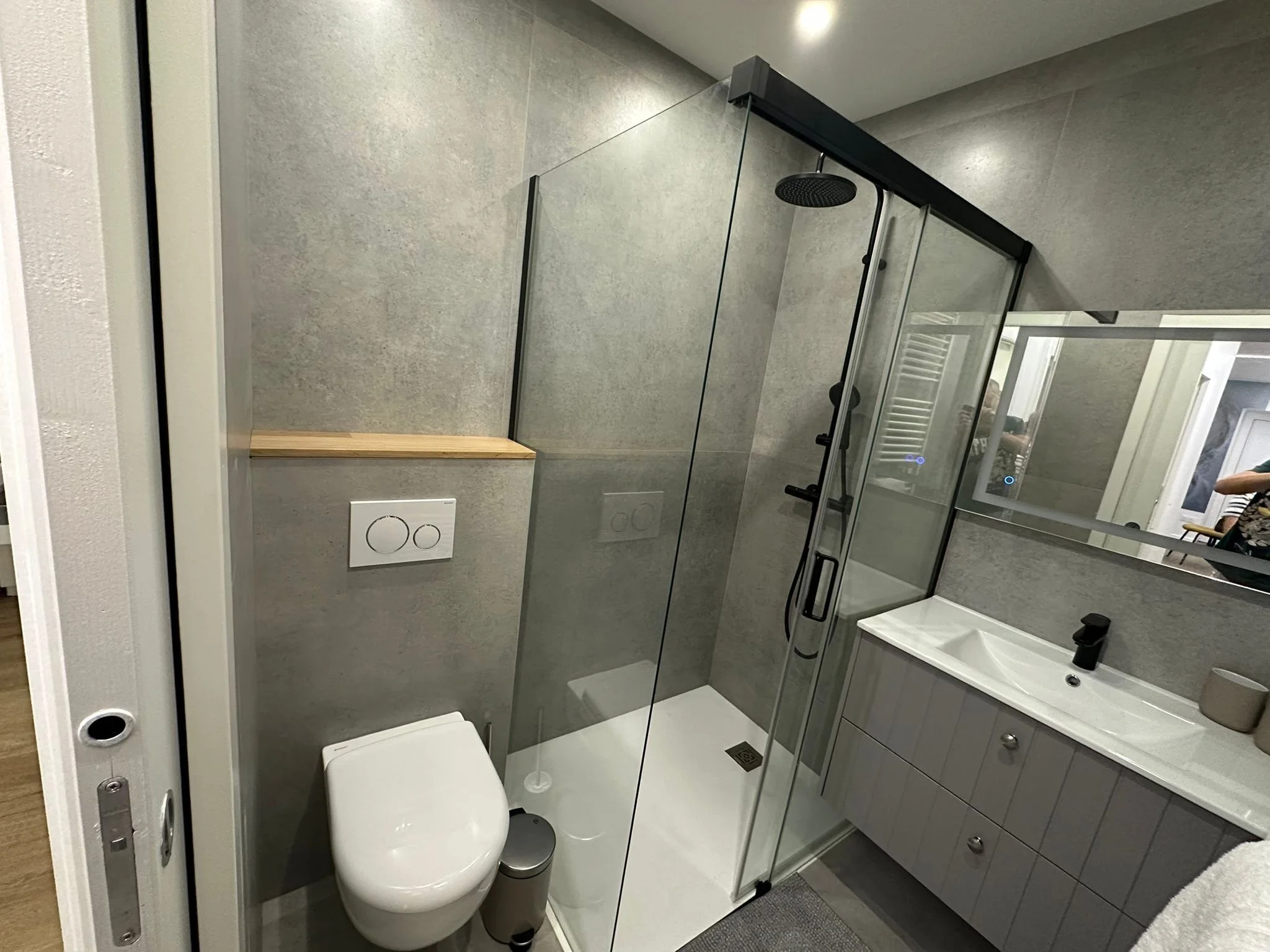 Une petite salle de bain moderne avec une cabine de douche en verre, un lavabo blanc avec tiroirs, un miroir au-dessus du lavabo, et un WC blanc. La décoration est épurée avec des murs gris et des appareils sanitaires intégrés.