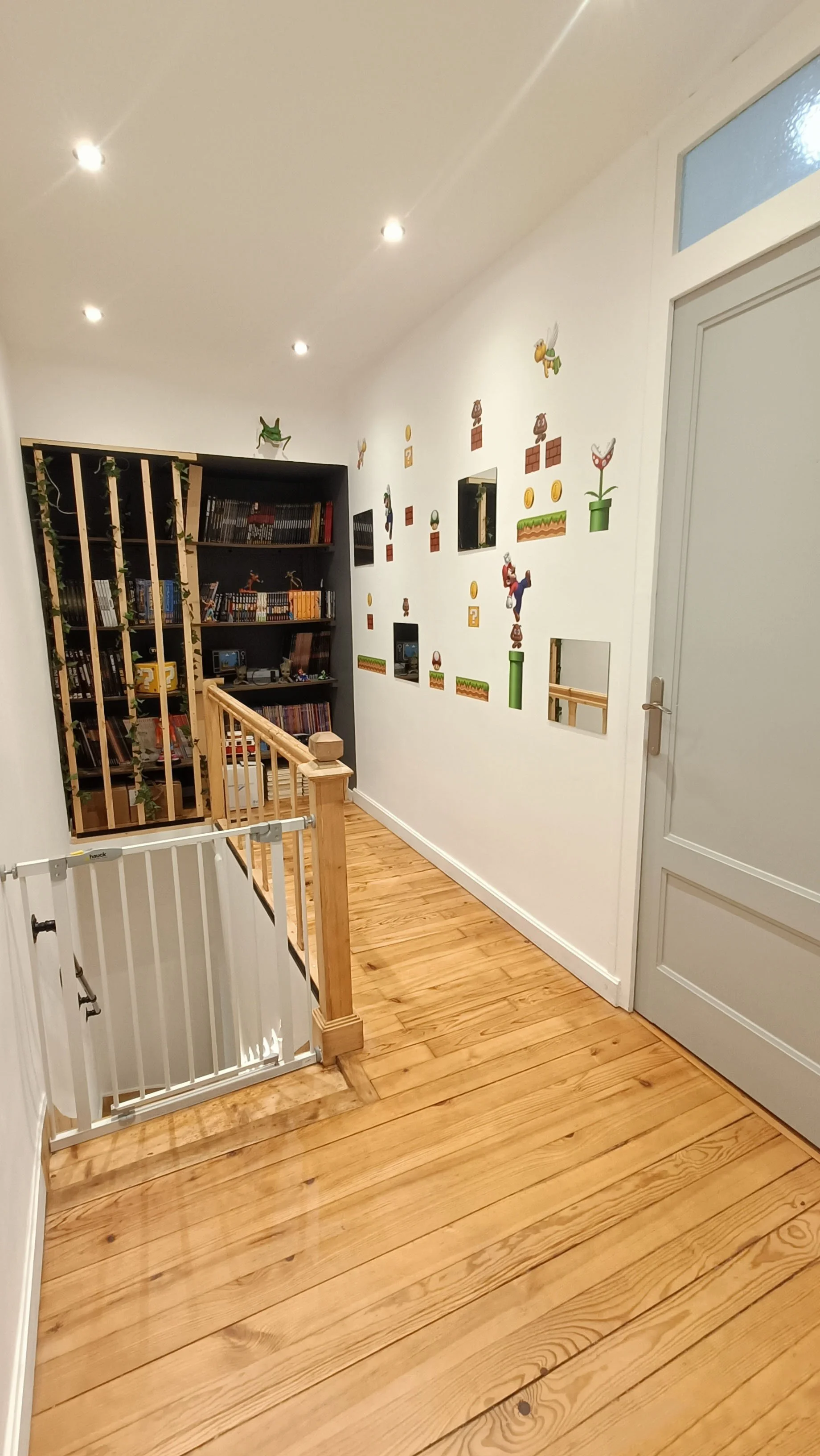 Un coin de couloir décoré avec des stickers de personnages de jeux vidéo sur le mur, un escabeau en bois et une bibliothèque remplie de livres et de figurines.