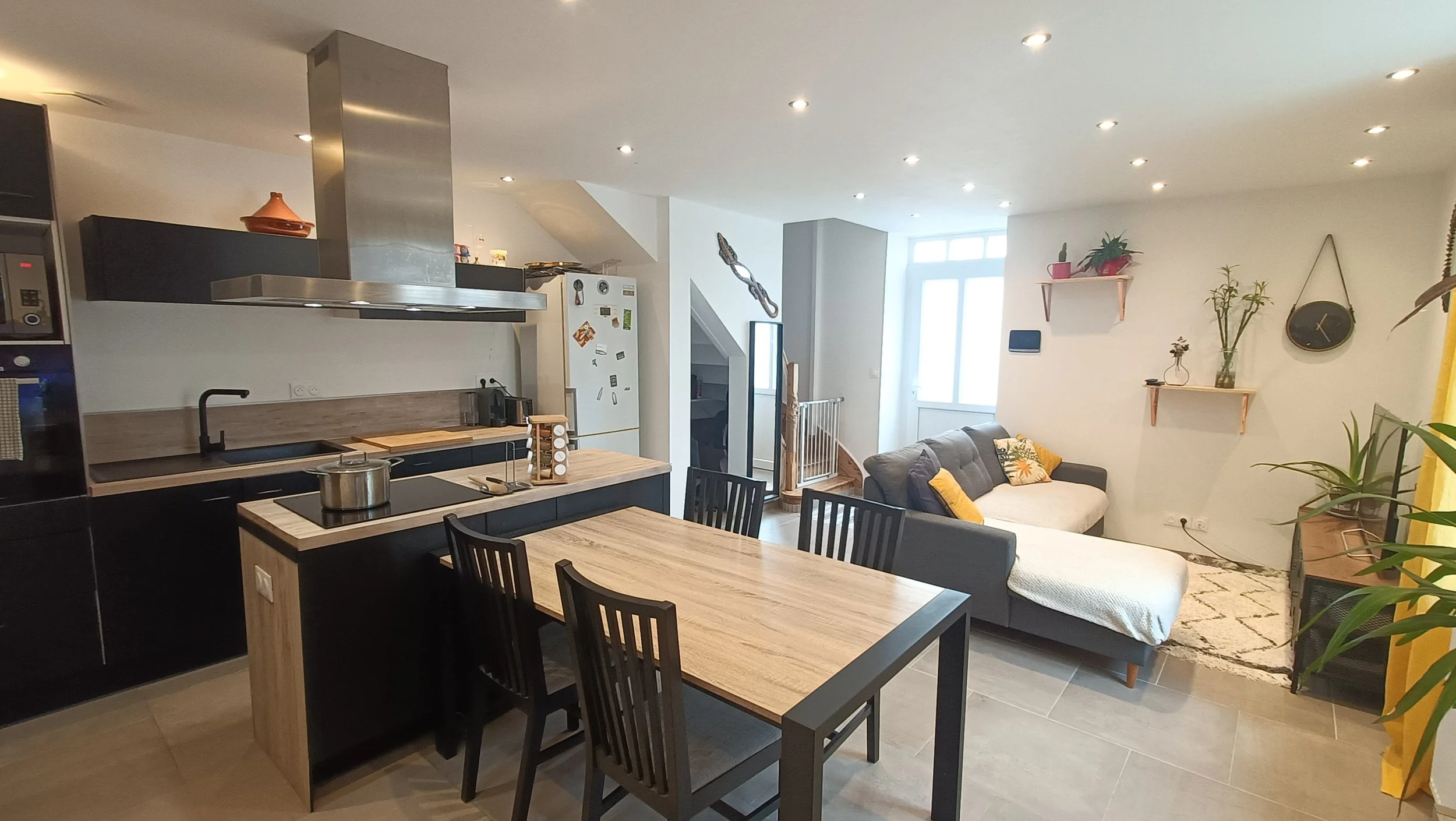 Intérieur d'un salon avec cuisine ouverte, comprenant un meuble de cuisine noir, une table à manger en bois, des chaises noires, un canapé gris avec des coussins jaunes, des étagères avec des plantes et une horloge ronde.