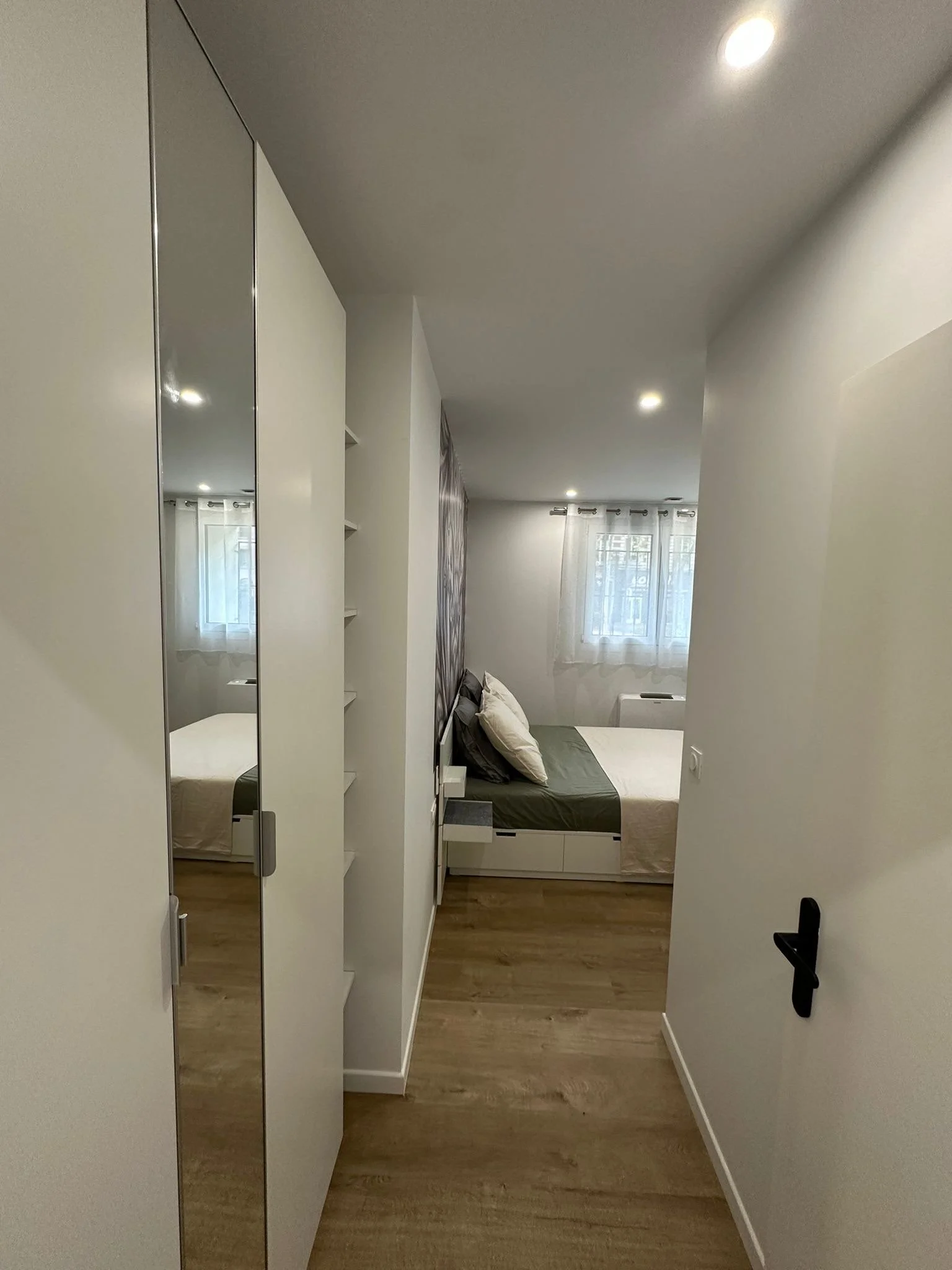 Une petite chambre de style moderne avec un lit, une tête de lit gris, une fenêtre avec des rideaux blancs, un placard avec miroir sur la porte, et un sol en bois.