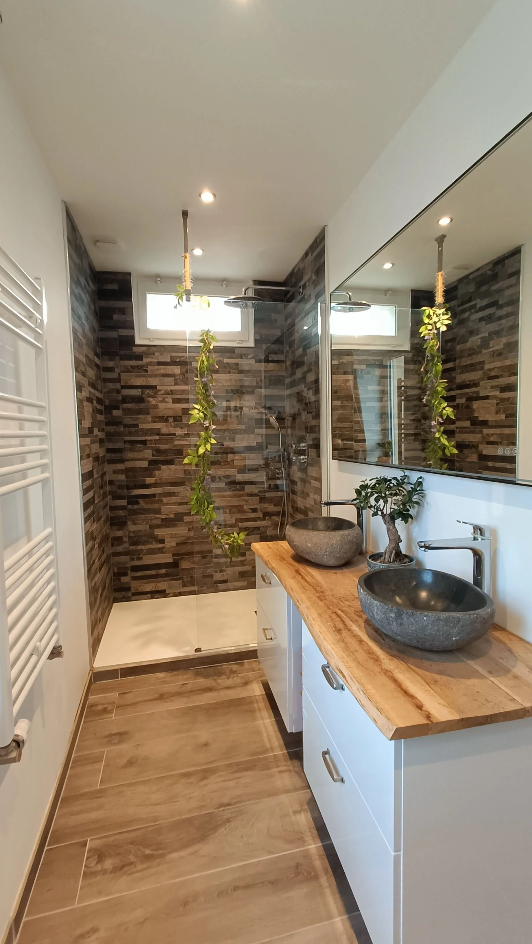 Une salle de bain moderne avec une douche en verre, murs recouverts de carreaux en pierre, une double vasque en pierre avec un plan en bois, miroirs et plantes décoratives.