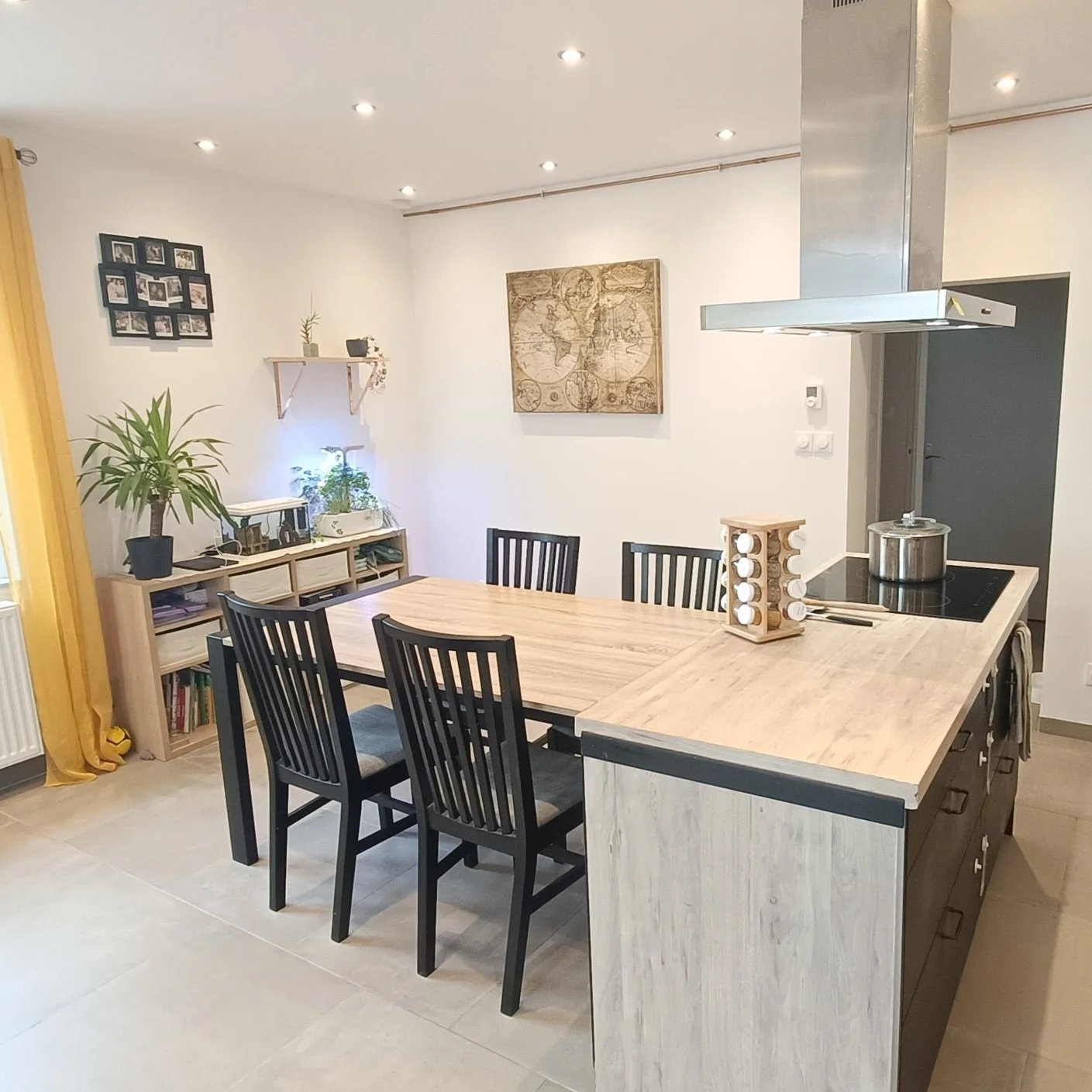 Cuisine moderne avec une table en bois clair entourée de six chaises noires, une cuisine équipée avec un îlot central, une hotte en acier inoxydable, des étagères en bois, des plantes, et des décorations murales, y compris une carte ancienne.