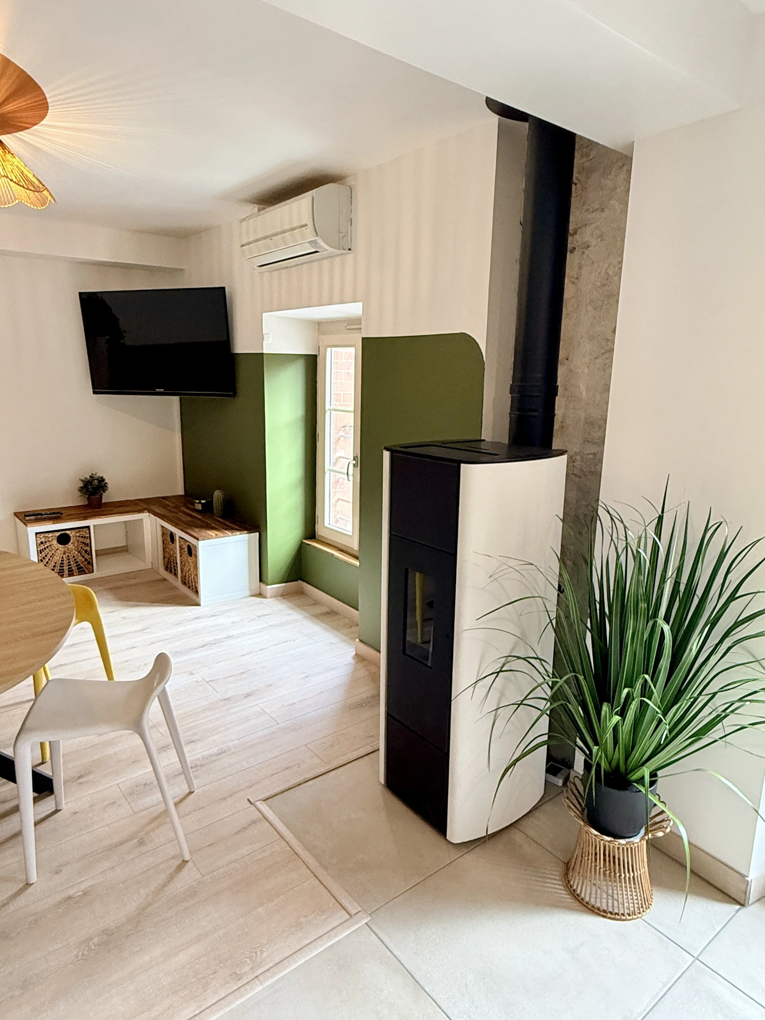 Intérieur d'une pièce avec un mur blanc et un mur vert, une télévision, une fenêtre, une cheminée électrique noire et blanche, une plante verte dans un pot noir sur un support en rotin, une table ronde avec des chaises.