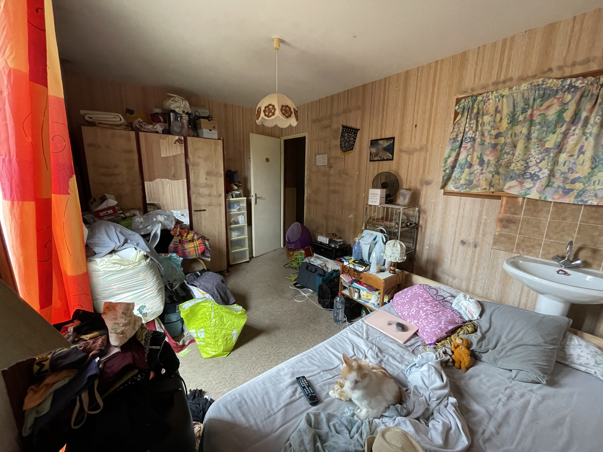 Intérieur en désordre d'une pièce avec des meubles, des vêtements, des objets divers, un chien posé sur un lit, une lampe suspendue et des rideaux fleuris.