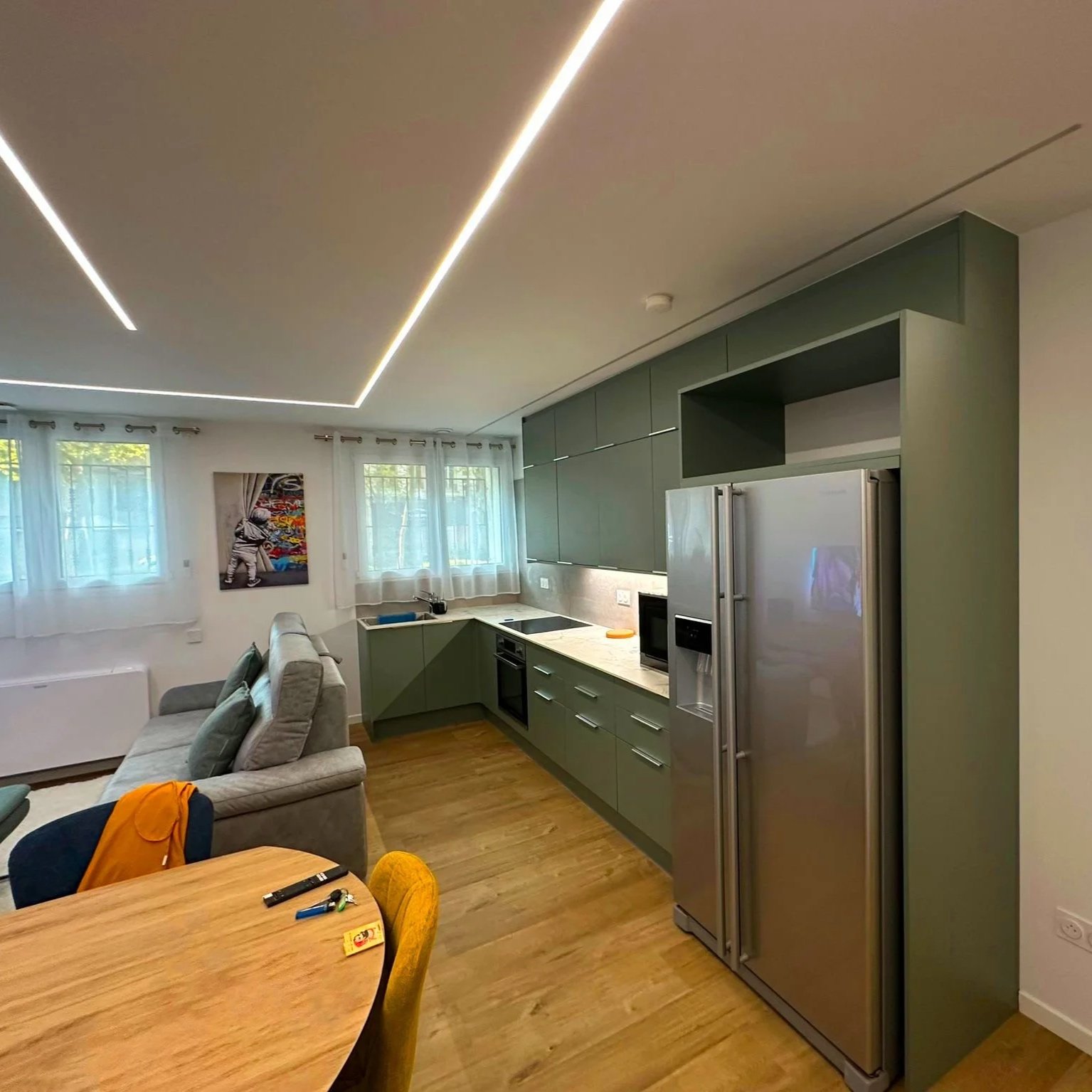 Une cuisine moderne avec des armoires vertes, un réfrigérateur en acier inoxydable, une table en bois, et un canapé gris visible dans le salon à côté.