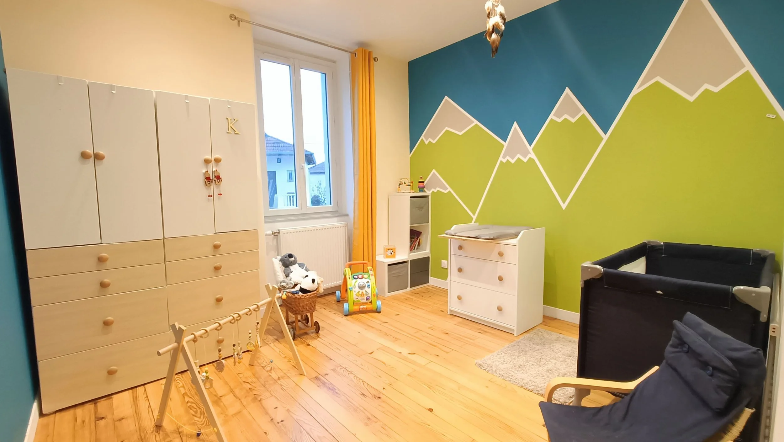 Une chambre d'enfant avec un mur peint de montagnes vertes, blanches et grises, contenant un lit en bois clair, une armoire blanche, un petit meuble blanc avec des jouets, un tapis beige, et une fenêtre avec des rideaux jaunes.