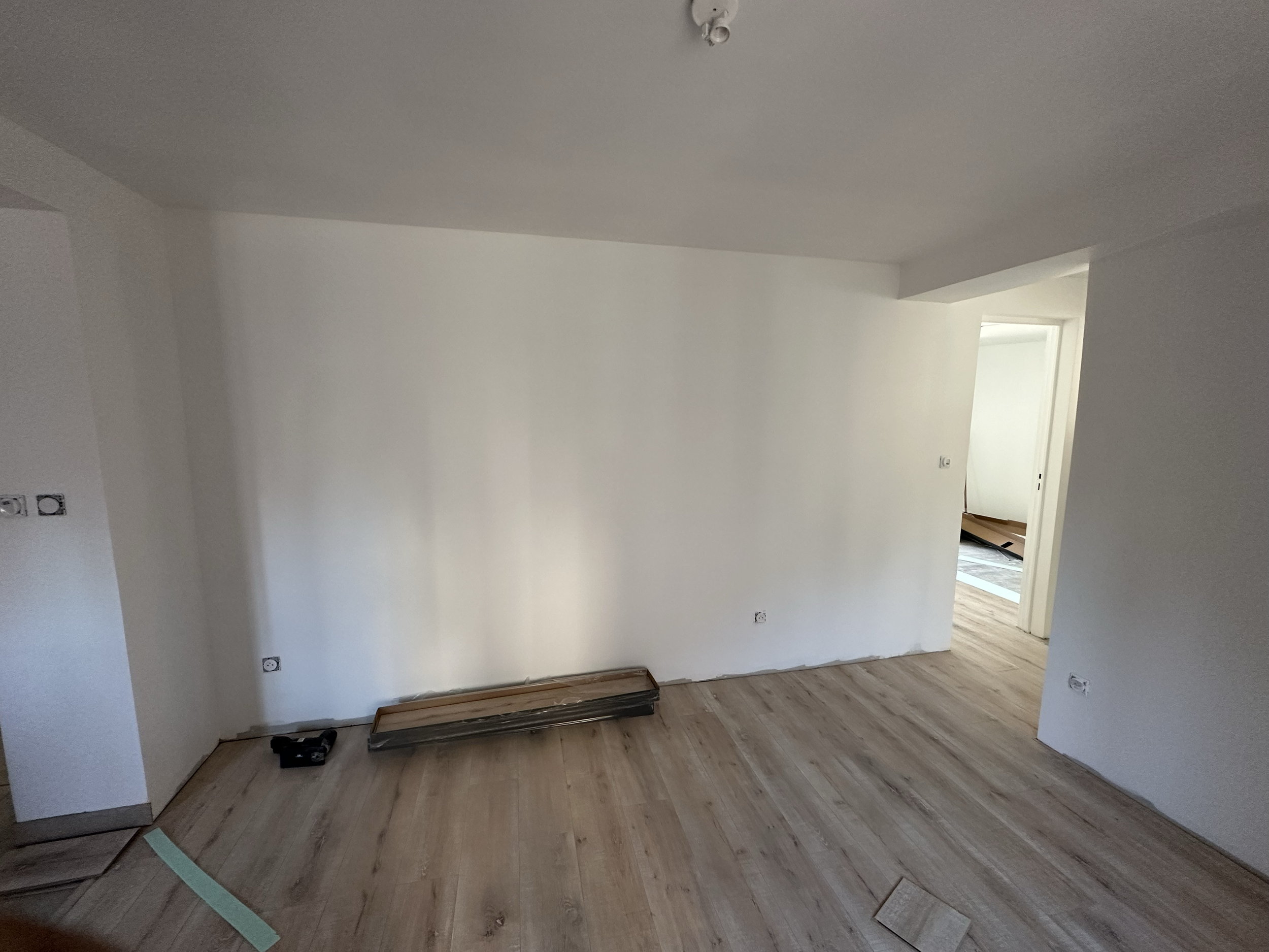 Salle en rénovation avec des murs blancs, un sol en bois clair, des prises électriques non installées, et un plafond avec un éclairage défectueux ou en cours de réparation.