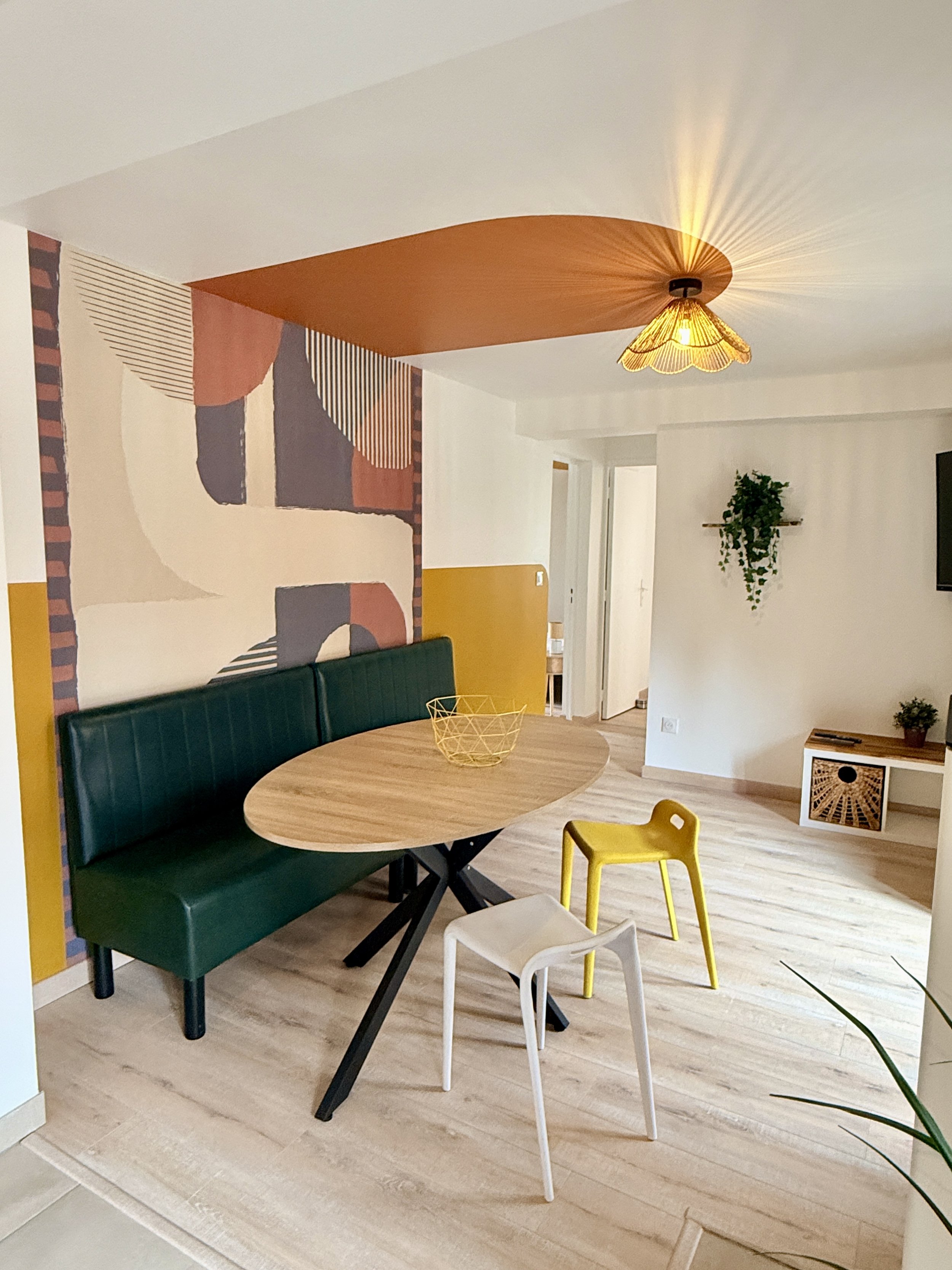 Salon moderne avec un mur mural abstrait, un plafond avec pendentif en rotin, un banc vert en cuir, une table en bois avec un panier en fil métallique, et plusieurs petites chaises colorées en blanc, jaune et beige. Il y a aussi des plantes décorativ