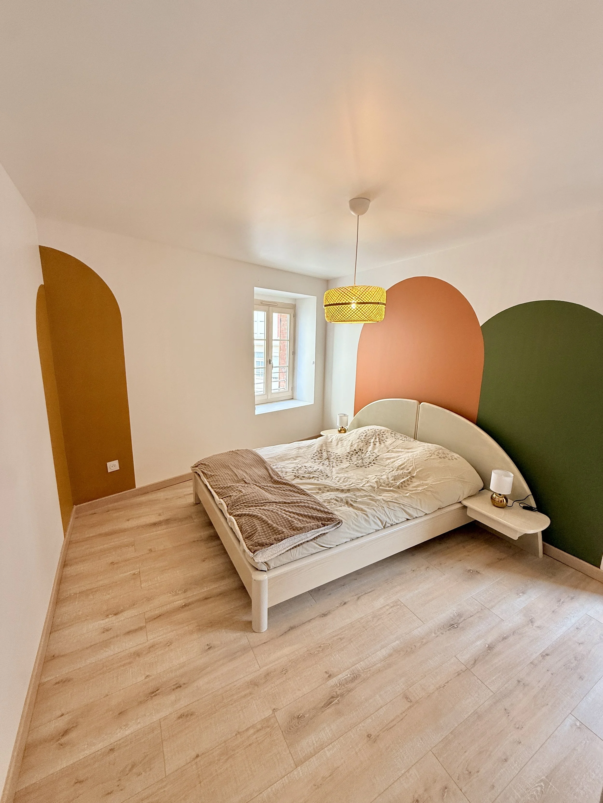 Une chambre moderne avec un lit double, deux lampes de chevet, un mur peint en deux couleurs (moutarde et vert foncé), une fenêtre laissant entrer la lumière naturelle, et un plafond avec un luminaire en rotin